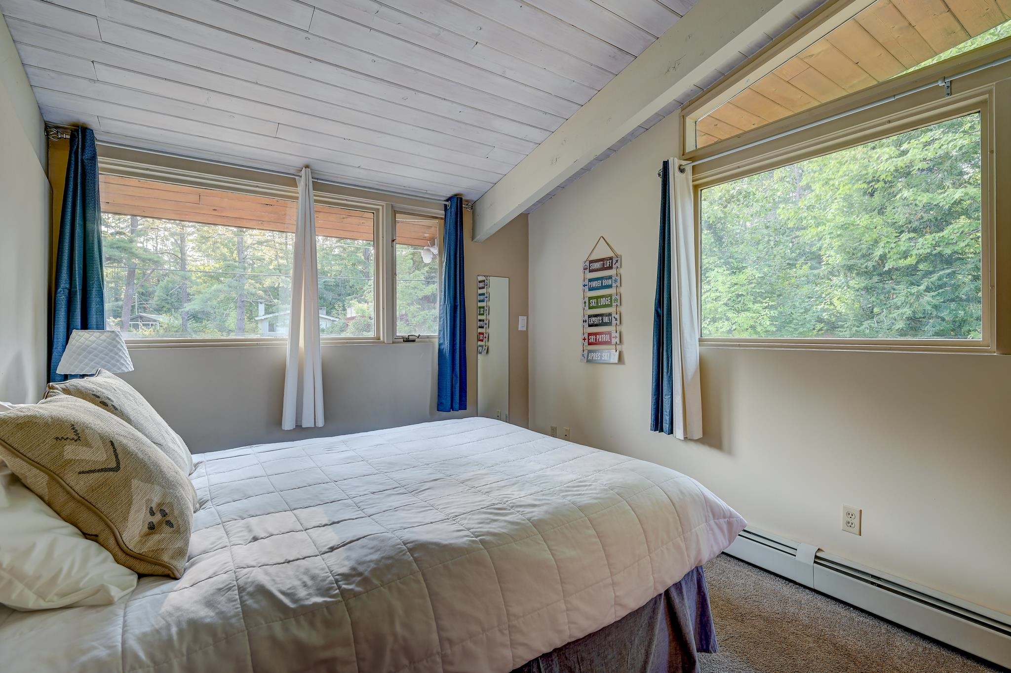 Bedroom with a white quilted bed, sloped plank ceiling, and large windows overlooking leafy trees.