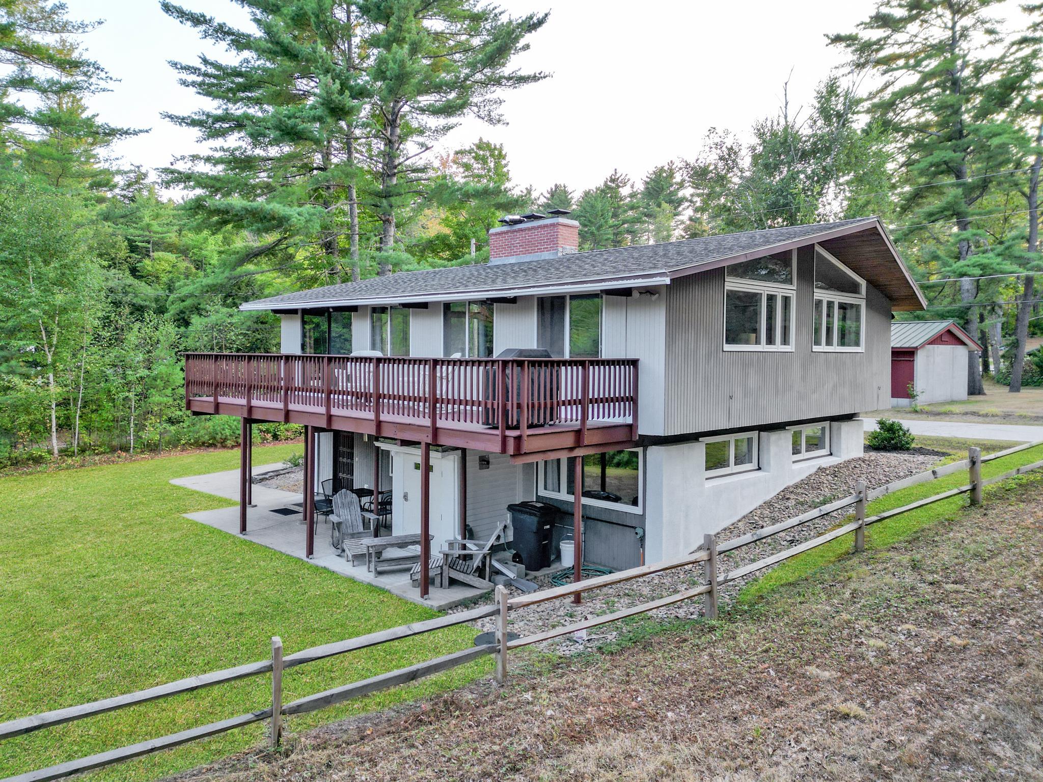 Raised split-level home with a long deck, lower patio, fenced lawn, and mature trees.
