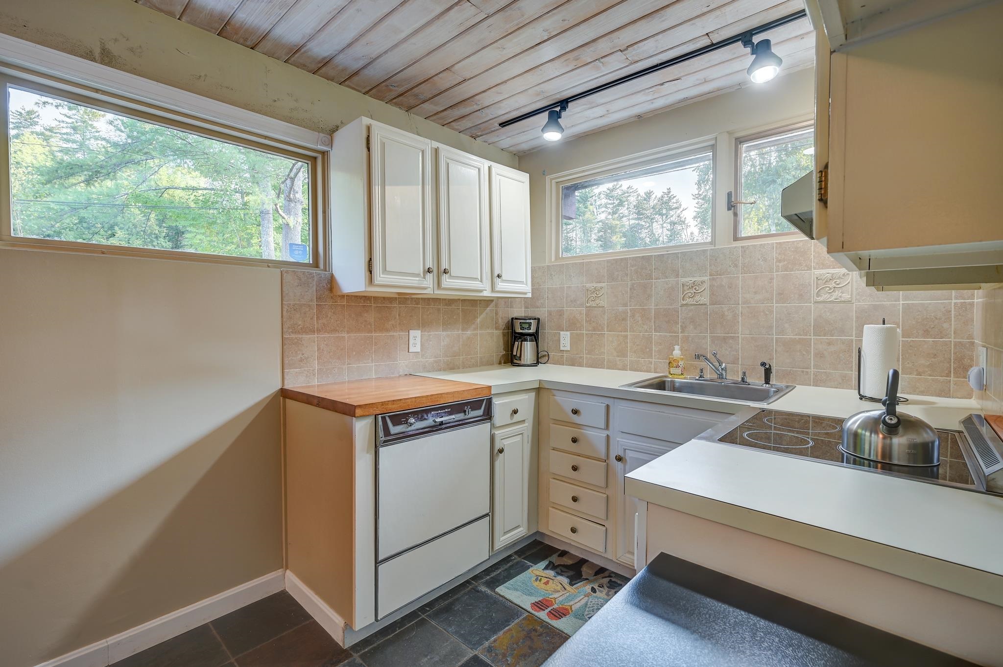 Kitchen with white cabinets, tiled backsplash, wood ceiling, and large windows overlooking trees.