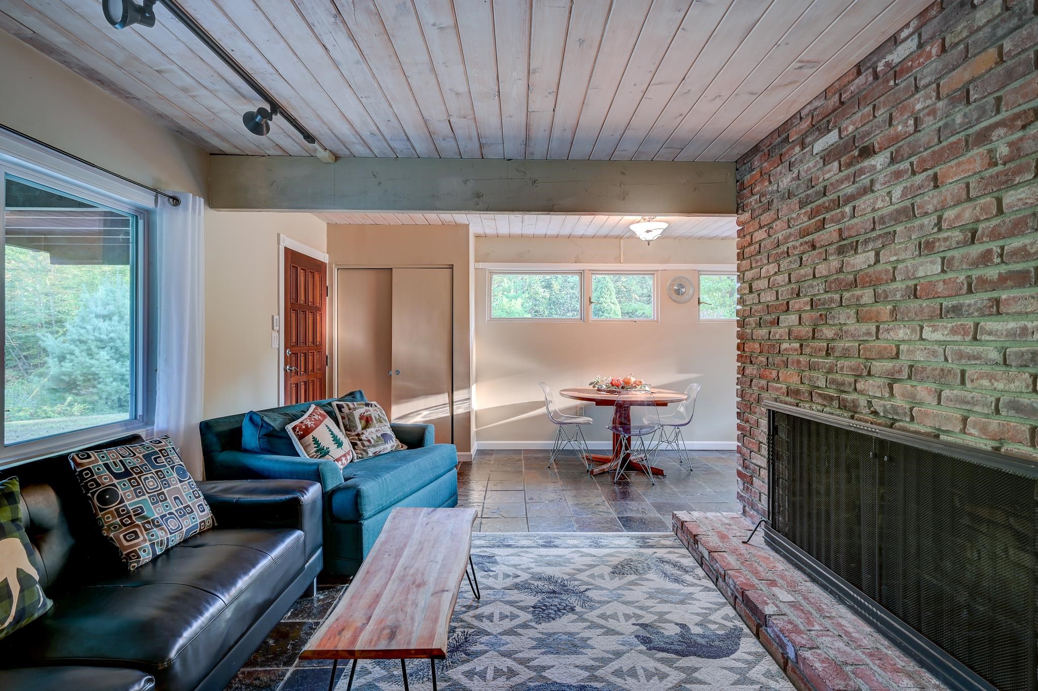 Living room with a brick fireplace, blue sofa, wood-plank ceiling, and adjoining dining area.