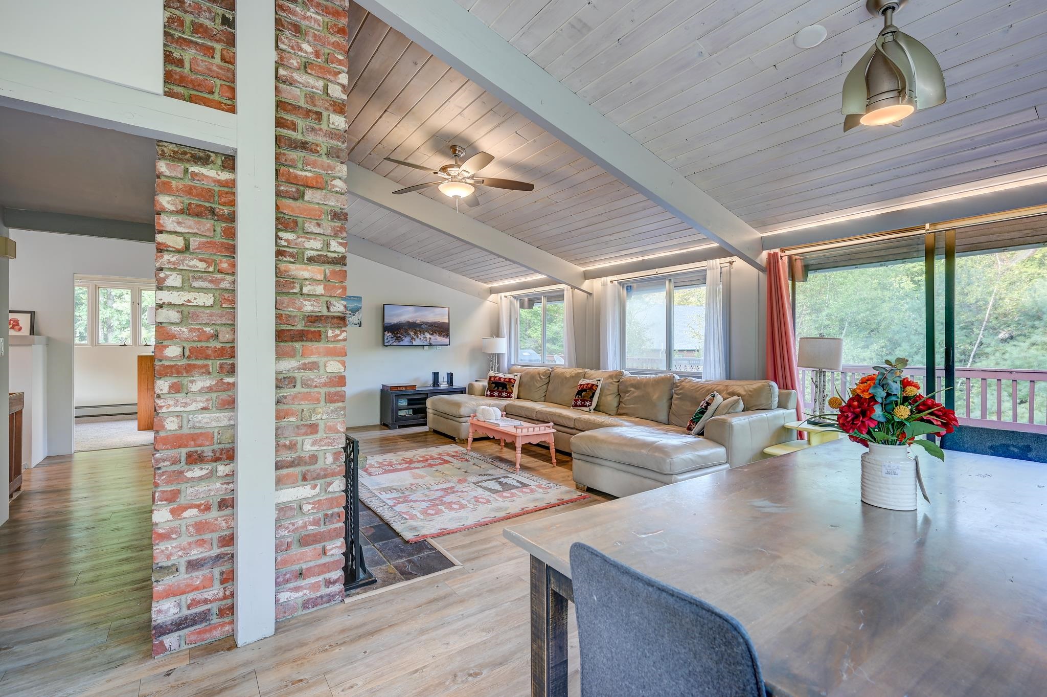 Living room with vaulted ceiling, brick columns, a beige sectional, and sliding doors overlooking trees.