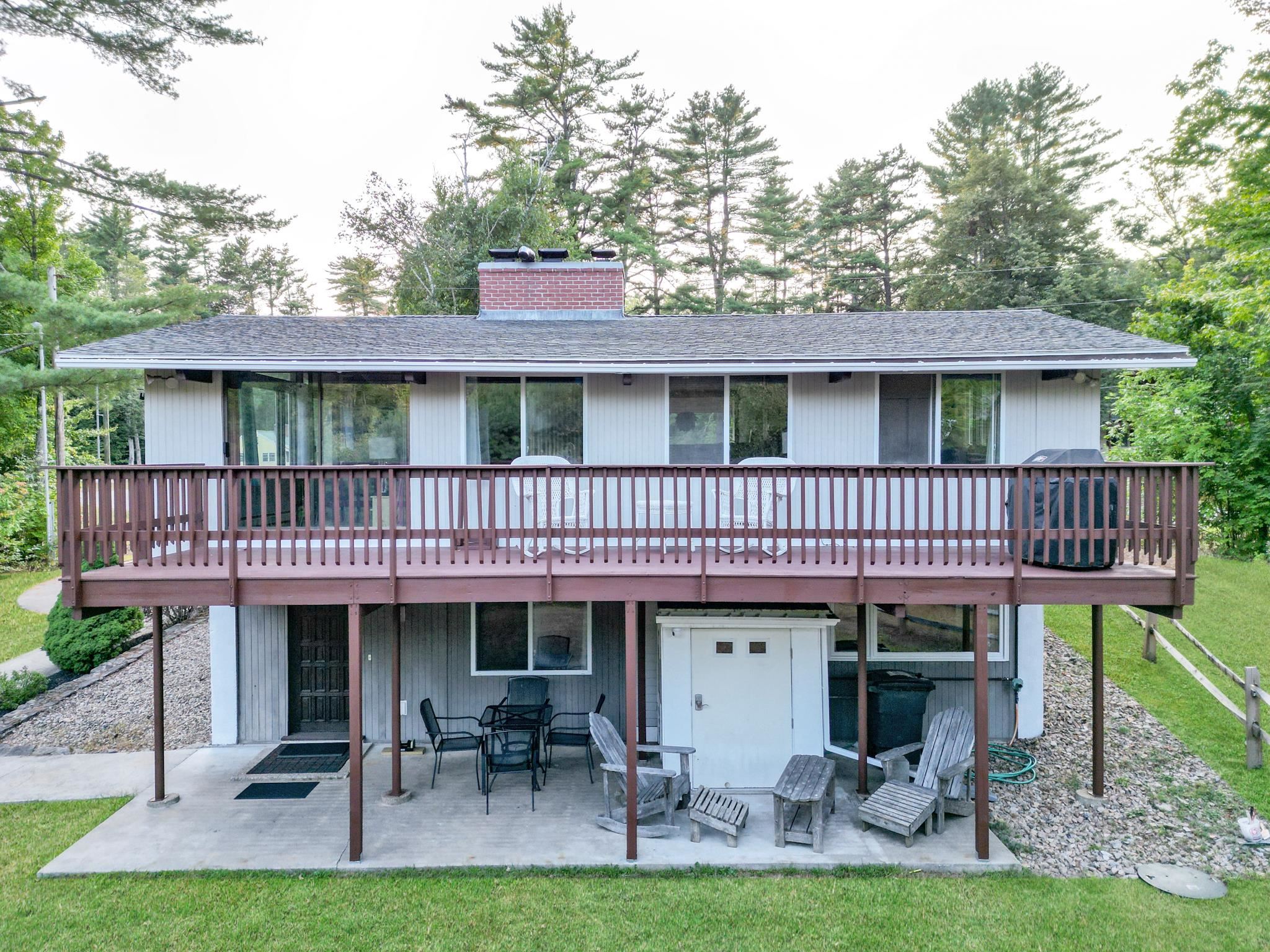 Two-story home with a wide upper deck, lower patio, and hot tub amid trees.