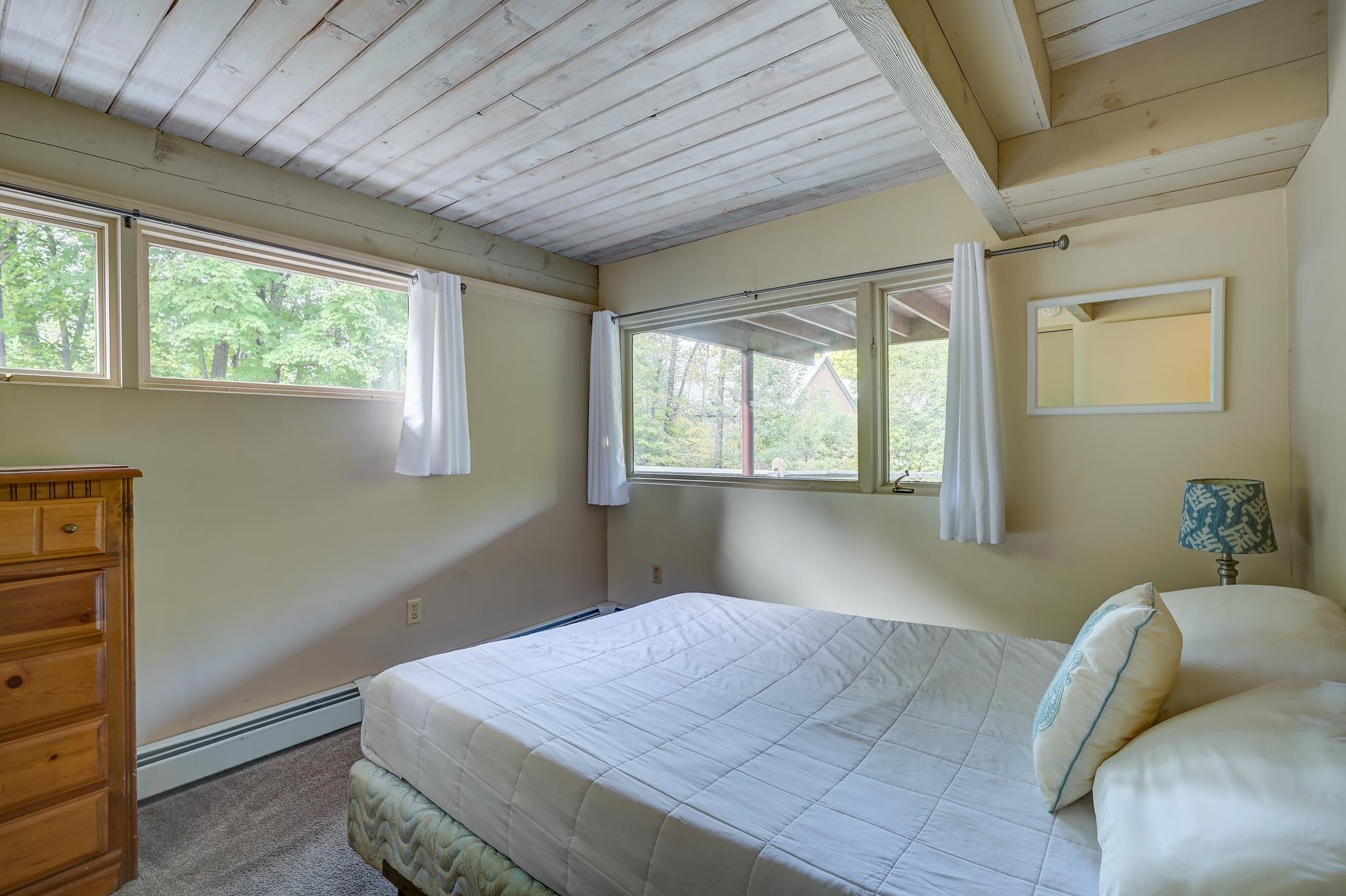 Bedroom with a white bed, wooden dresser, and windows overlooking leafy trees beneath a white plank ceiling.