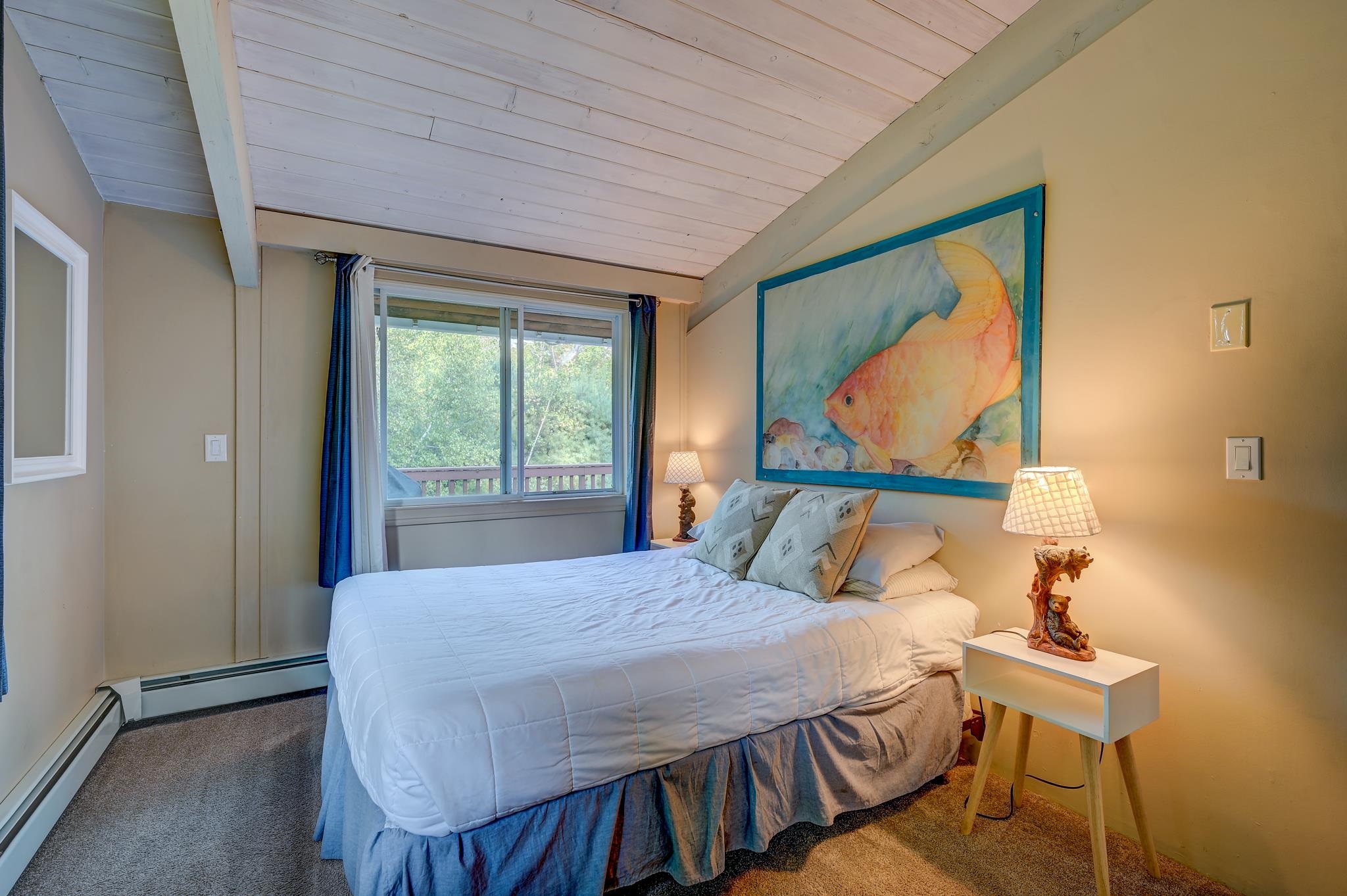 Bedroom with a vaulted plank ceiling, large fish painting, and window overlooking trees.