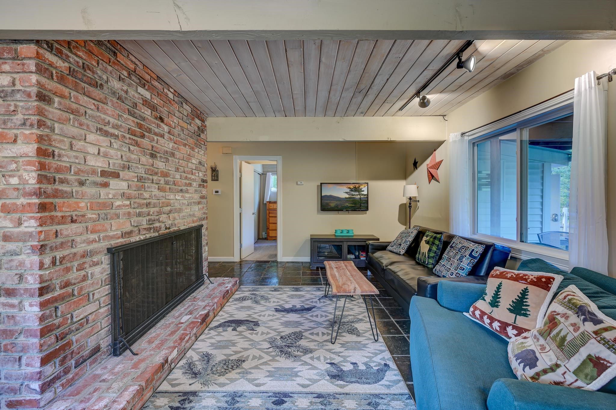 Living room with a brick fireplace, blue sofas, wide windows, and a wood-plank ceiling.