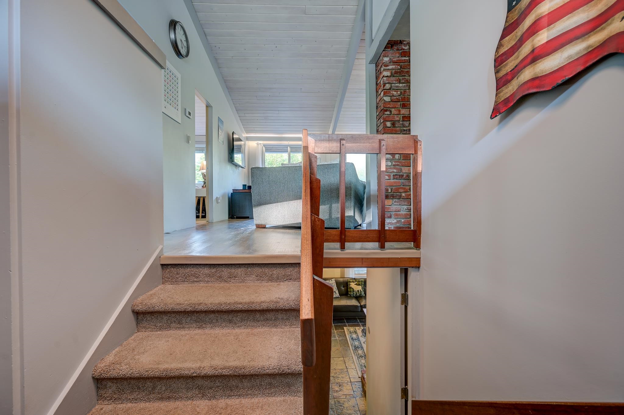 Carpeted stairs lead to a bright living area with a wood-and-glass railing and exposed brick column.