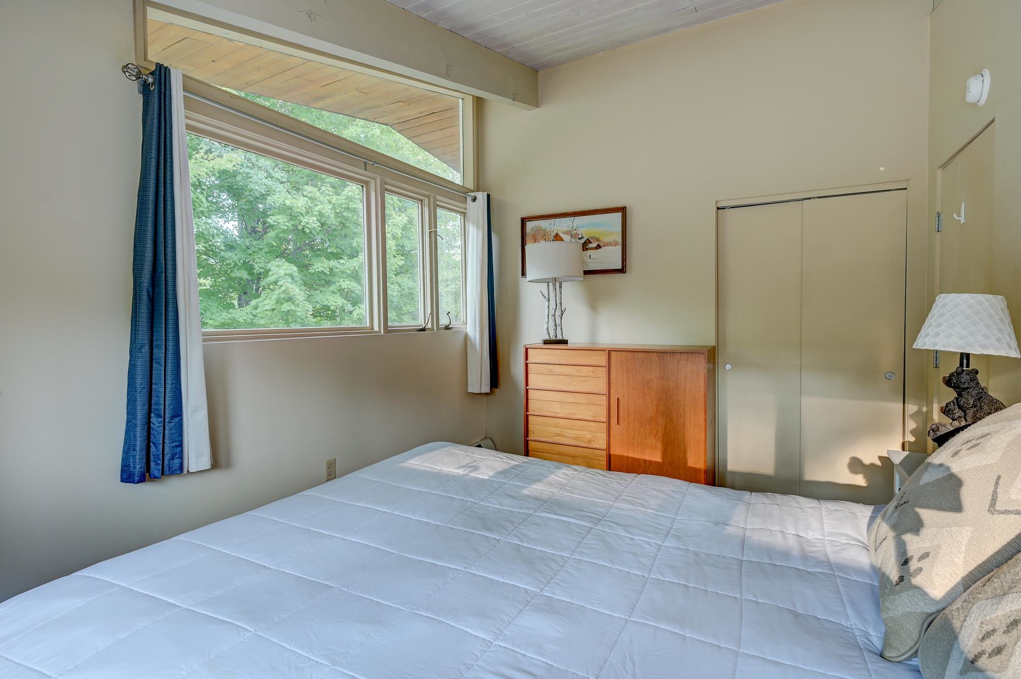 Bedroom with a white bed, large tree-view windows, blue curtains, and a wooden dresser.