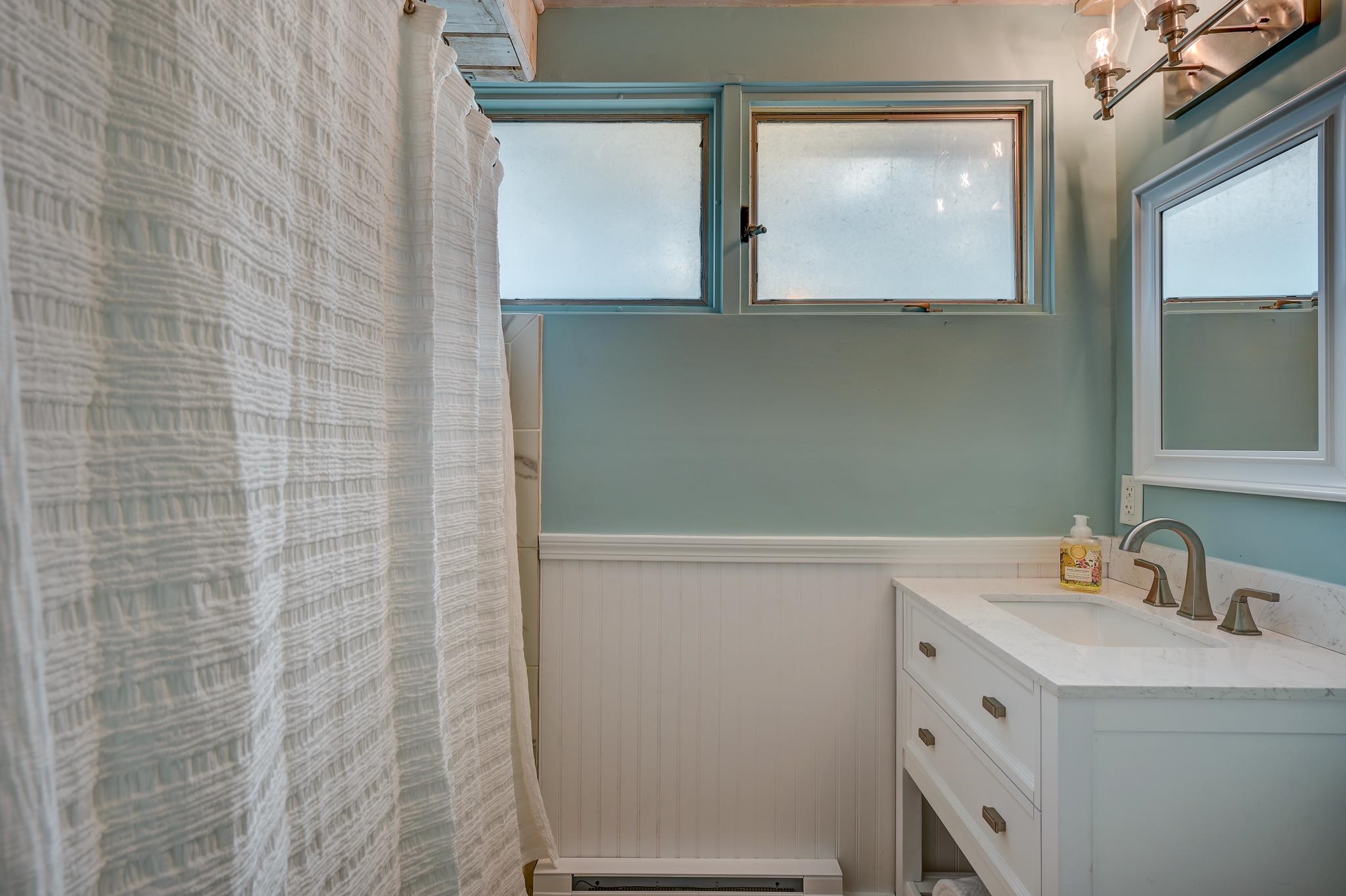 Bathroom with a textured white shower curtain, pale blue-green walls, and a white vanity beneath a mirror.