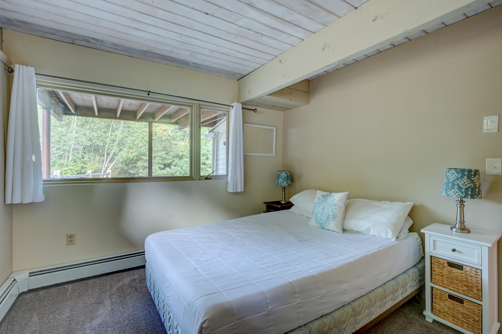 Bedroom with a white bed beneath a sloped plank ceiling and a wide window overlooking trees.