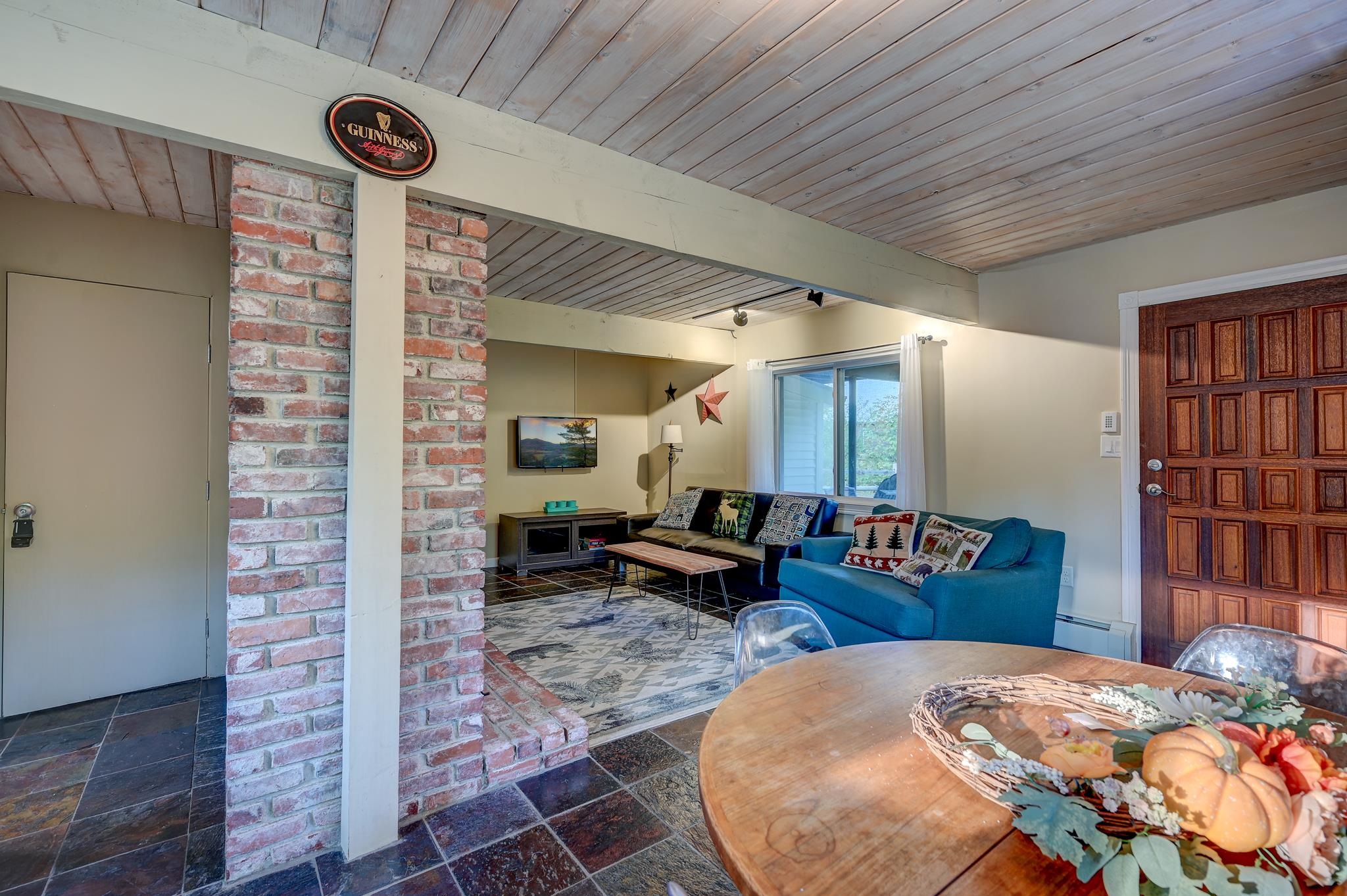 Living and dining area with a blue sofa, exposed brick column, and wood-plank ceiling.