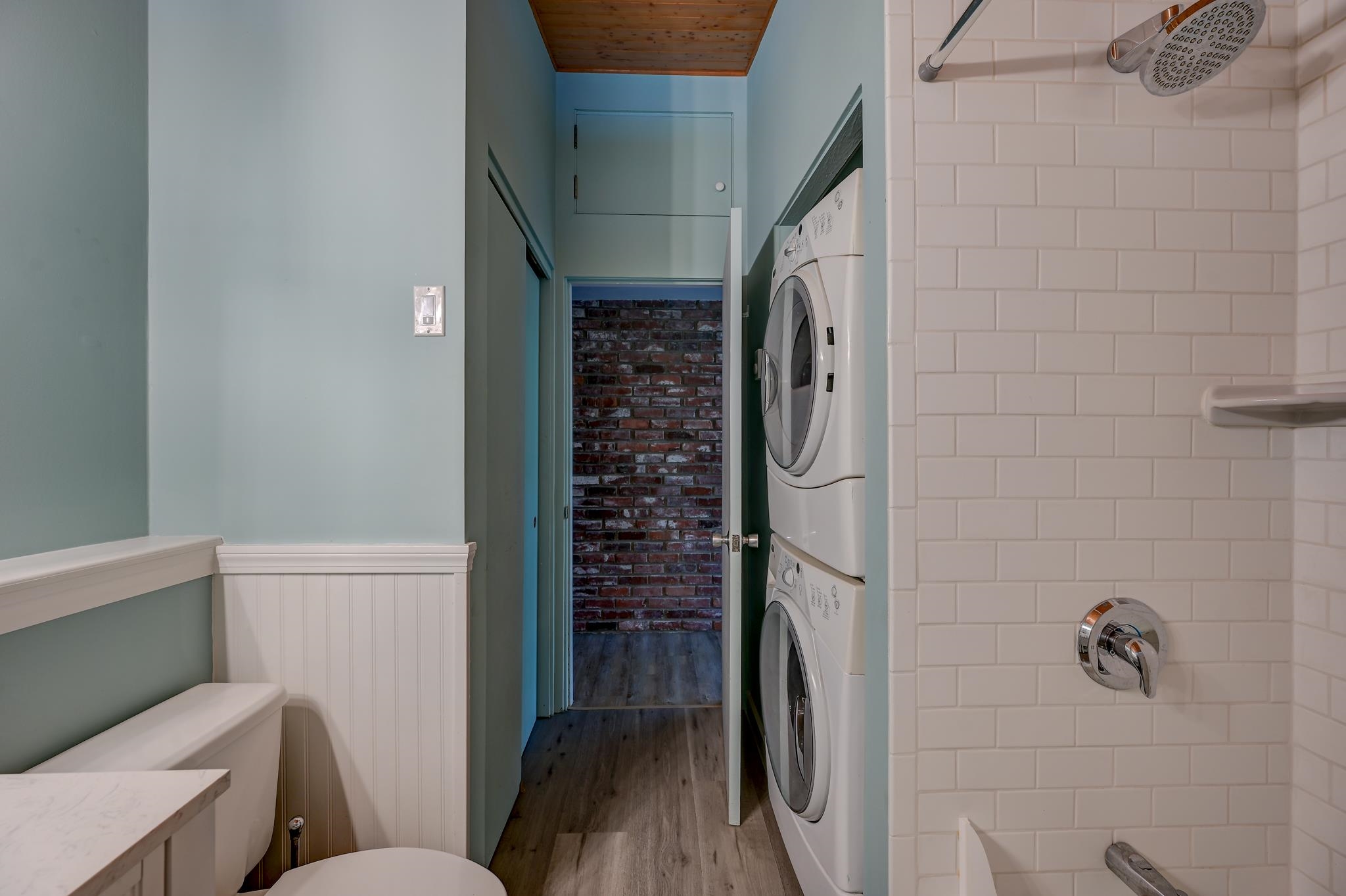 Bathroom with a tiled shower and toilet, next to a hallway with stacked washer and dryer.