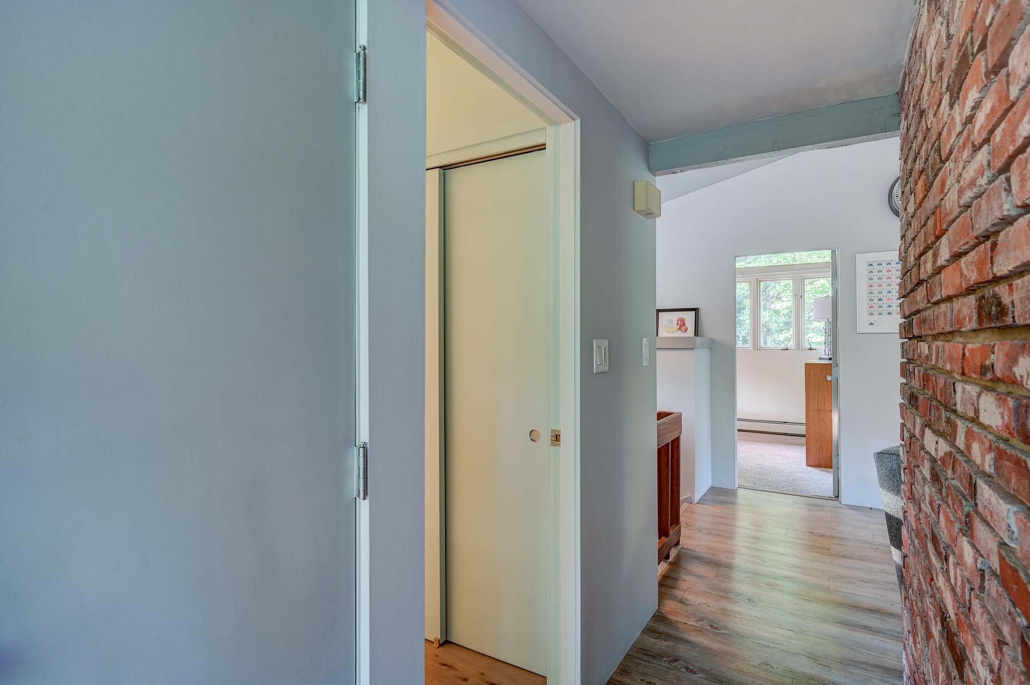 Hallway with an exposed brick wall and wood-plank flooring, opening into a bright room with windows.