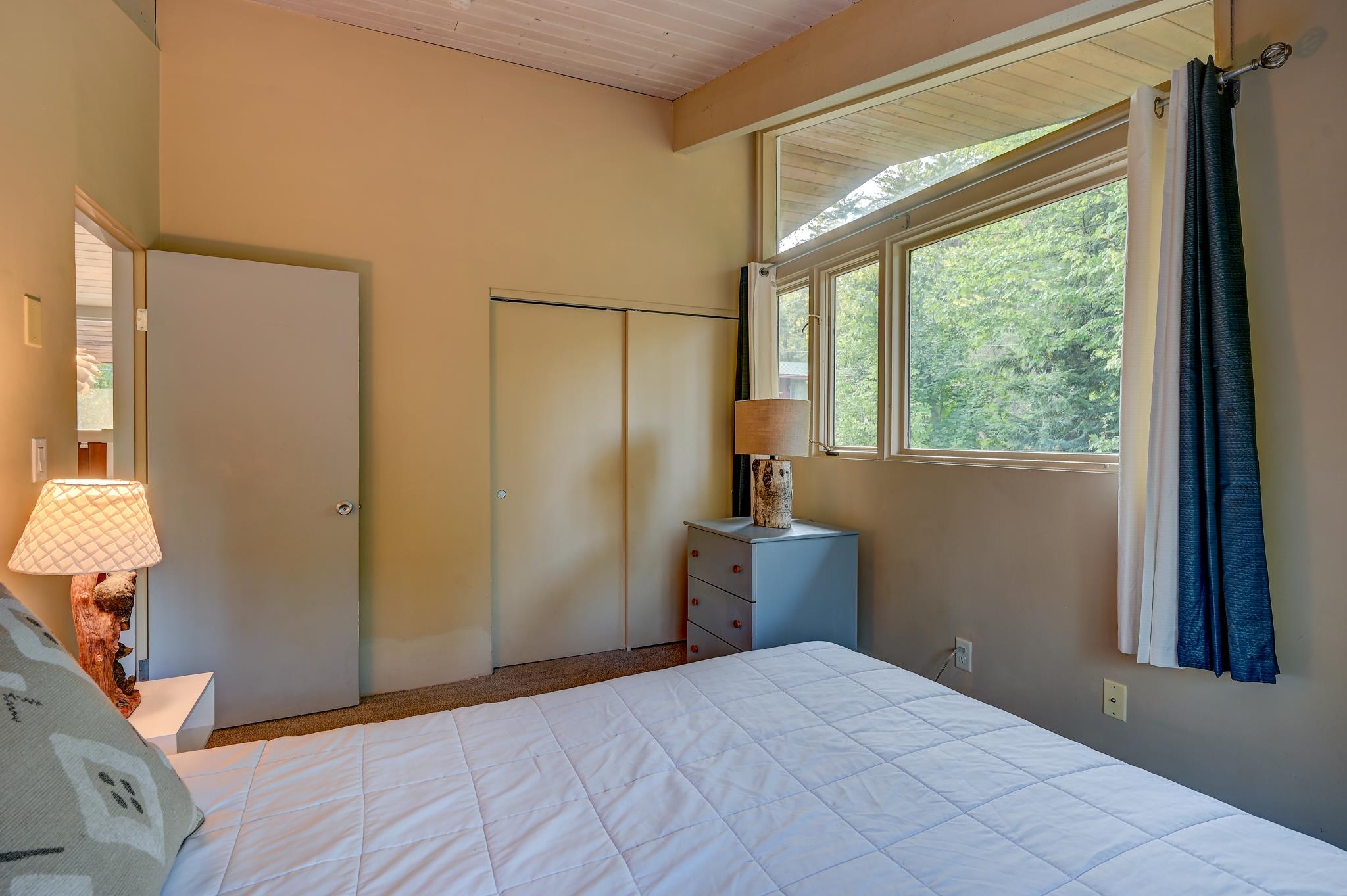 Bedroom with a white quilted bed, vaulted wood ceiling, and large windows overlooking leafy trees.
