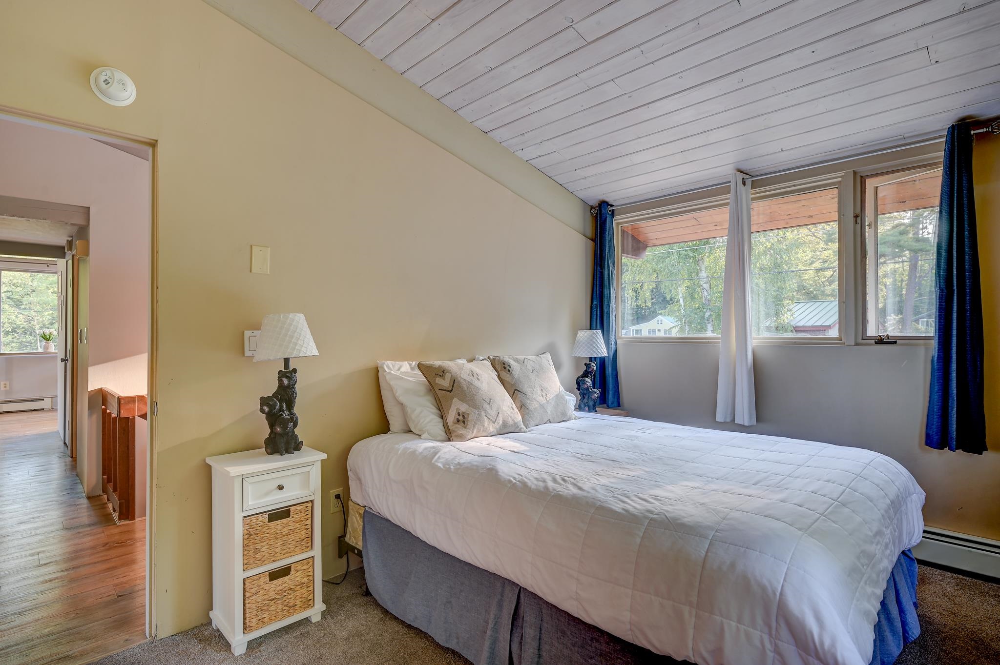 Bedroom with a white-covered bed, sloped plank ceiling, and large windows overlooking trees.