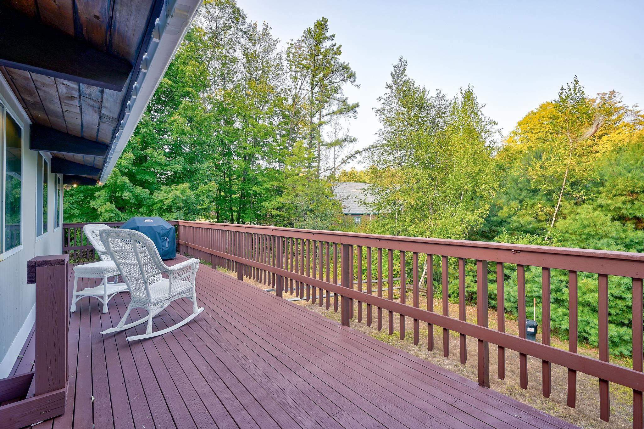 Raised deck with white wicker rocking chairs and a covered grill, overlooking trees in early autumn.