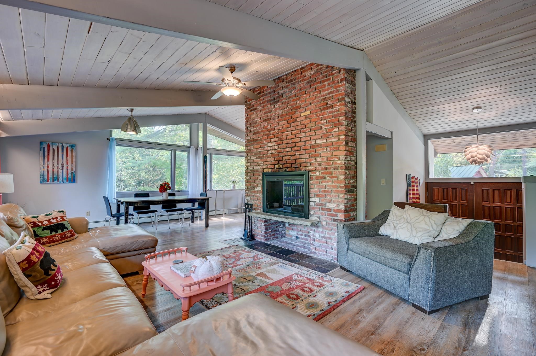Open-plan living room with a tall brick fireplace, vaulted ceiling, and large windows beside the dining area.