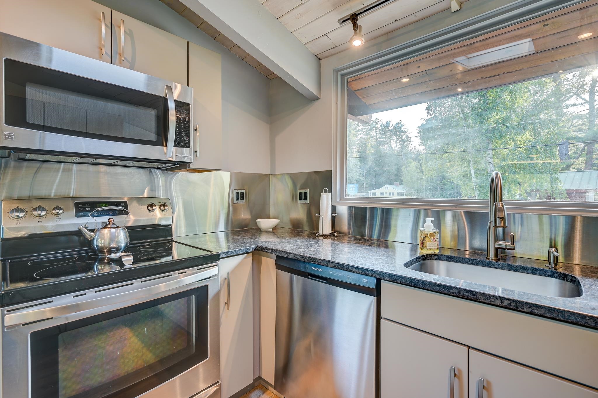 Modern kitchen with stainless steel appliances and a sink beneath a large window overlooking trees.