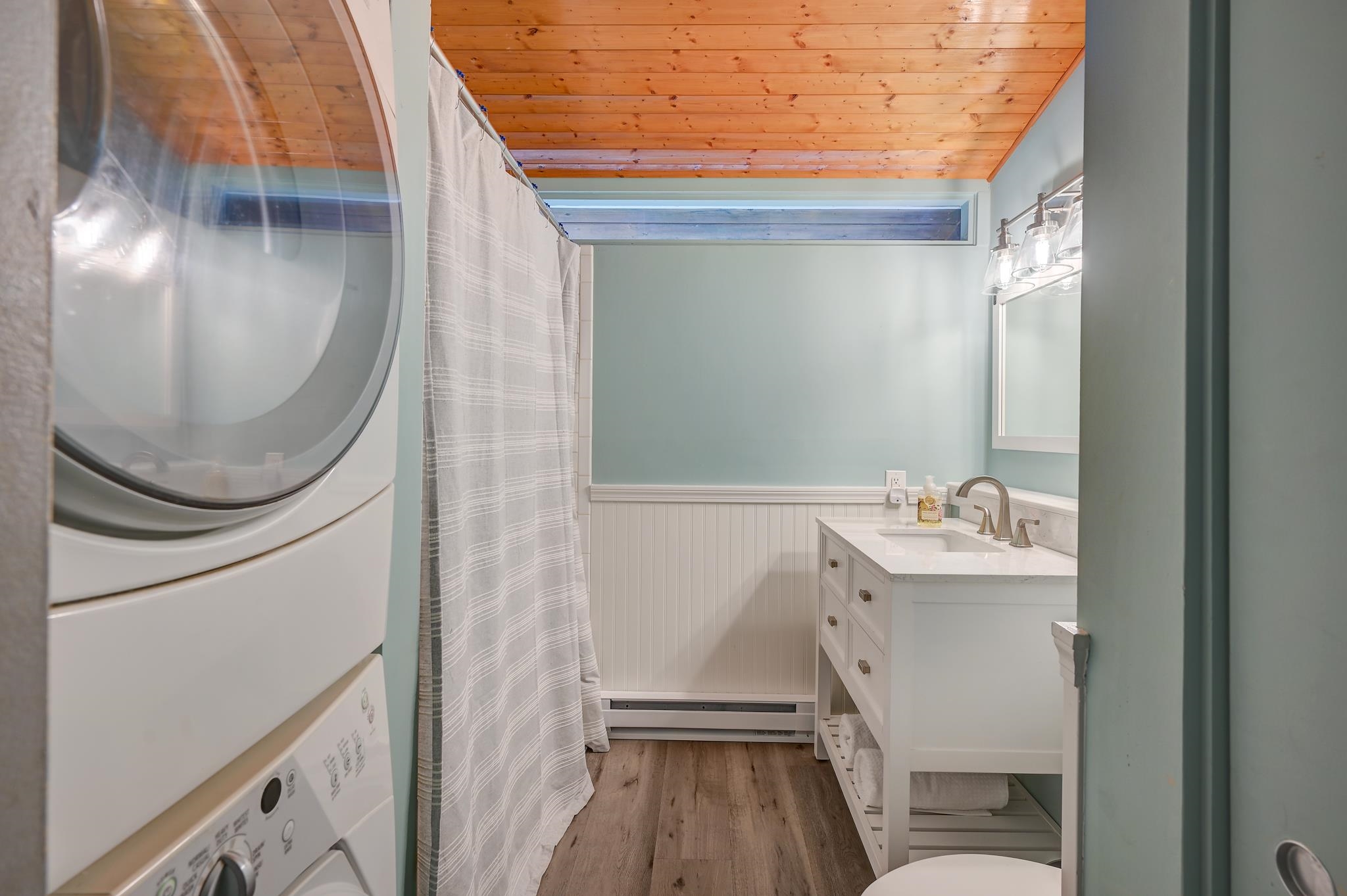 Bathroom with stacked washer and dryer, curtained shower, white vanity, and wood-plank ceiling.