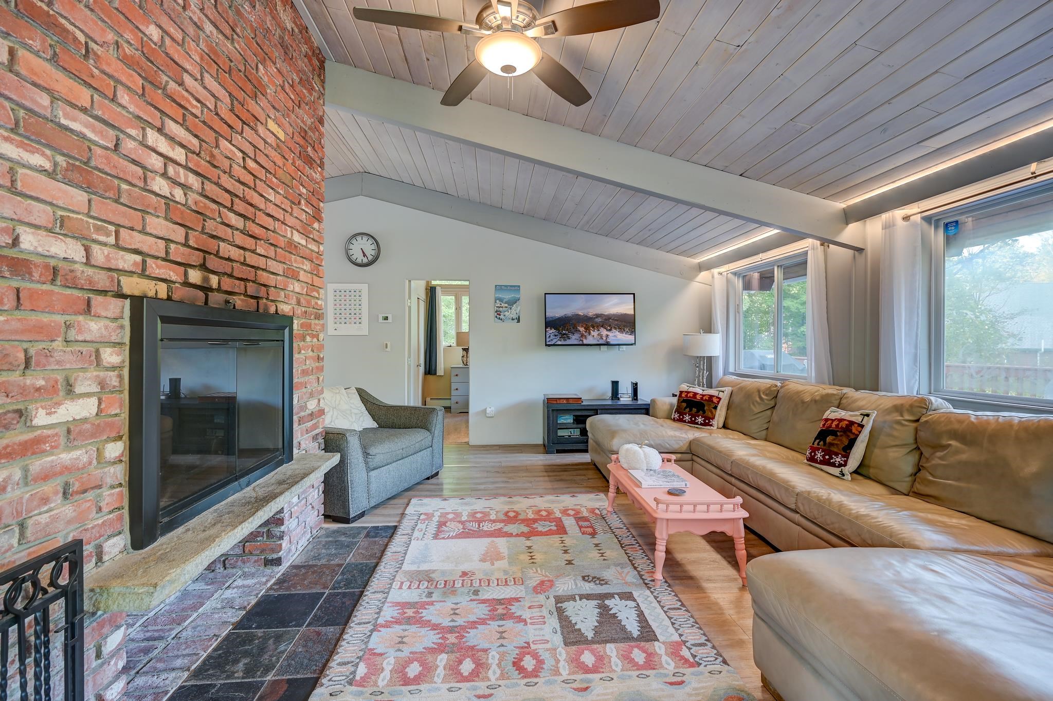 Living room with a brick fireplace, vaulted ceiling, leather sectional, and large windows.