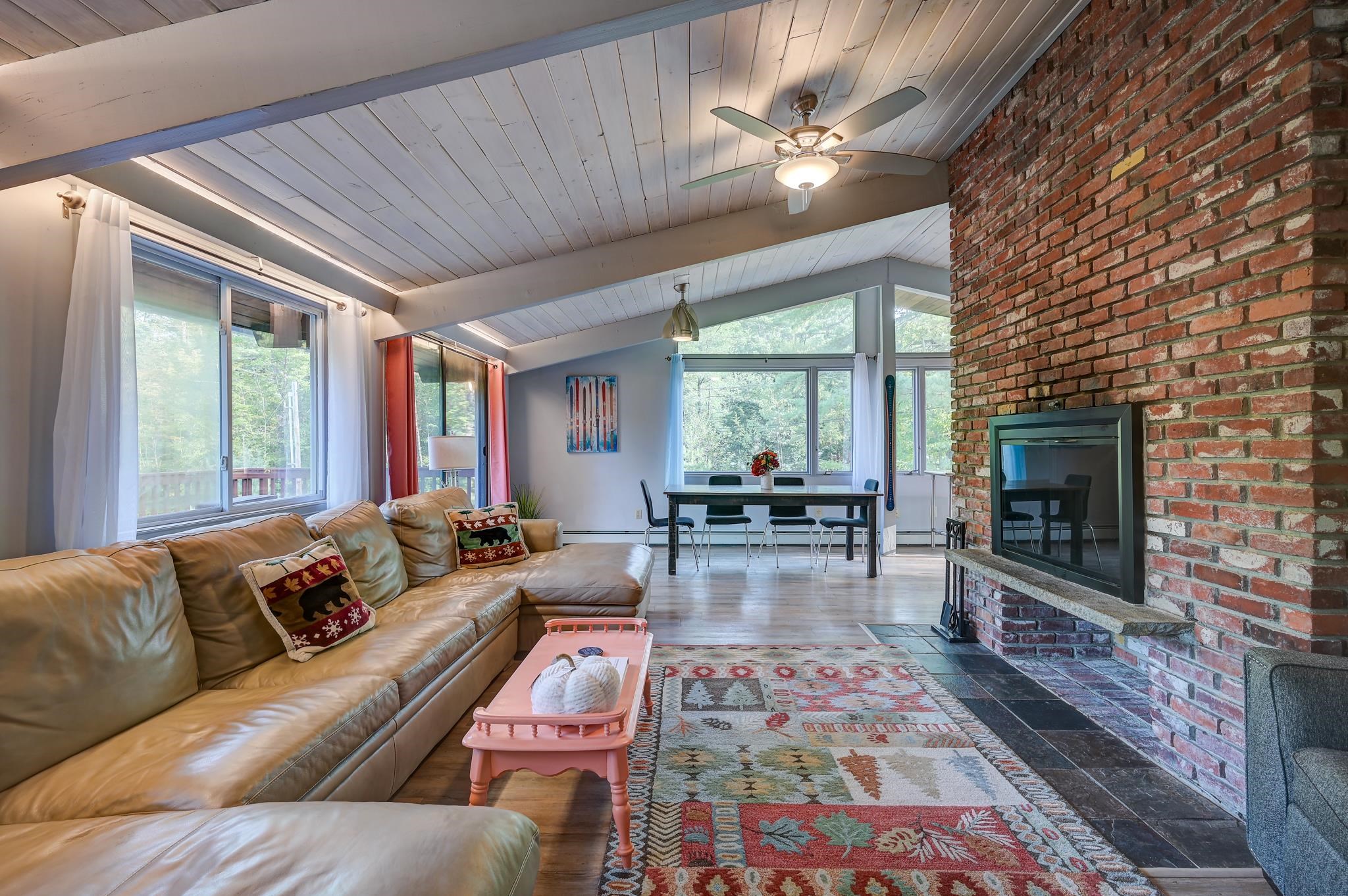 Open-plan living and dining room with a vaulted ceiling, leather sectional, large windows, and brick fireplace.