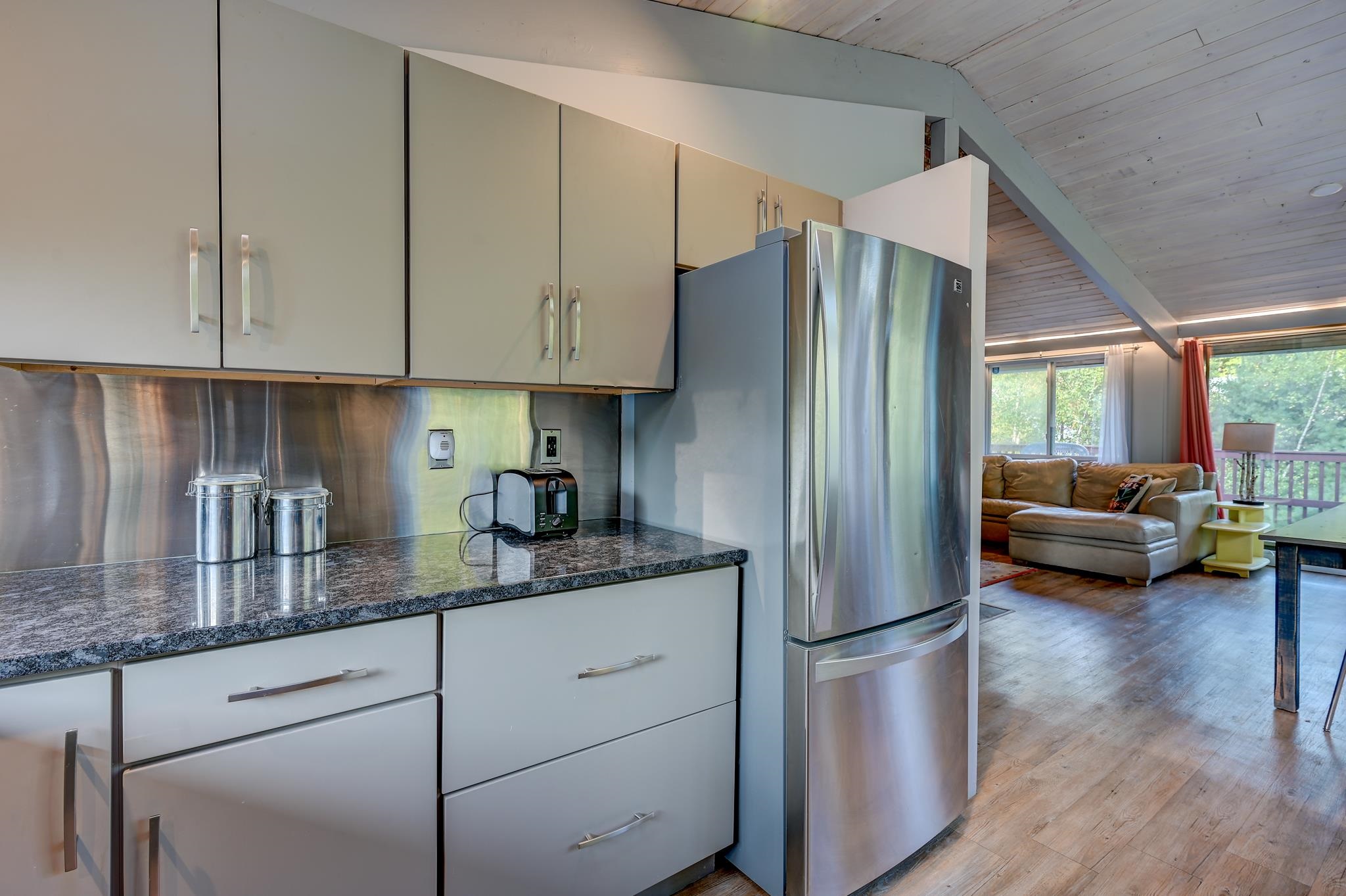 Kitchen with granite counters and stainless refrigerator opening to a vaulted living area and deck.