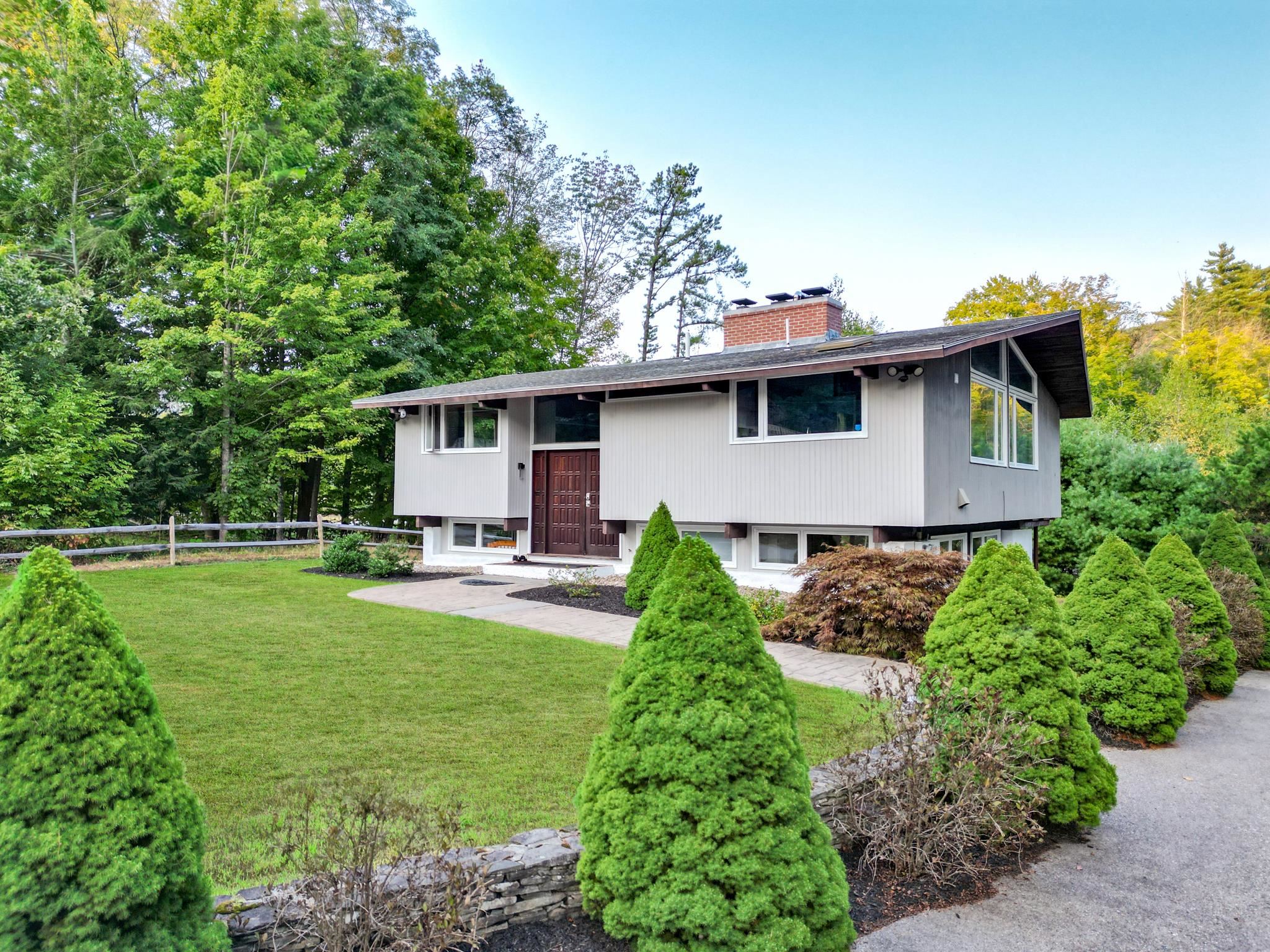 Split-level home with broad windows, a landscaped lawn, clipped evergreens, and mature trees.
