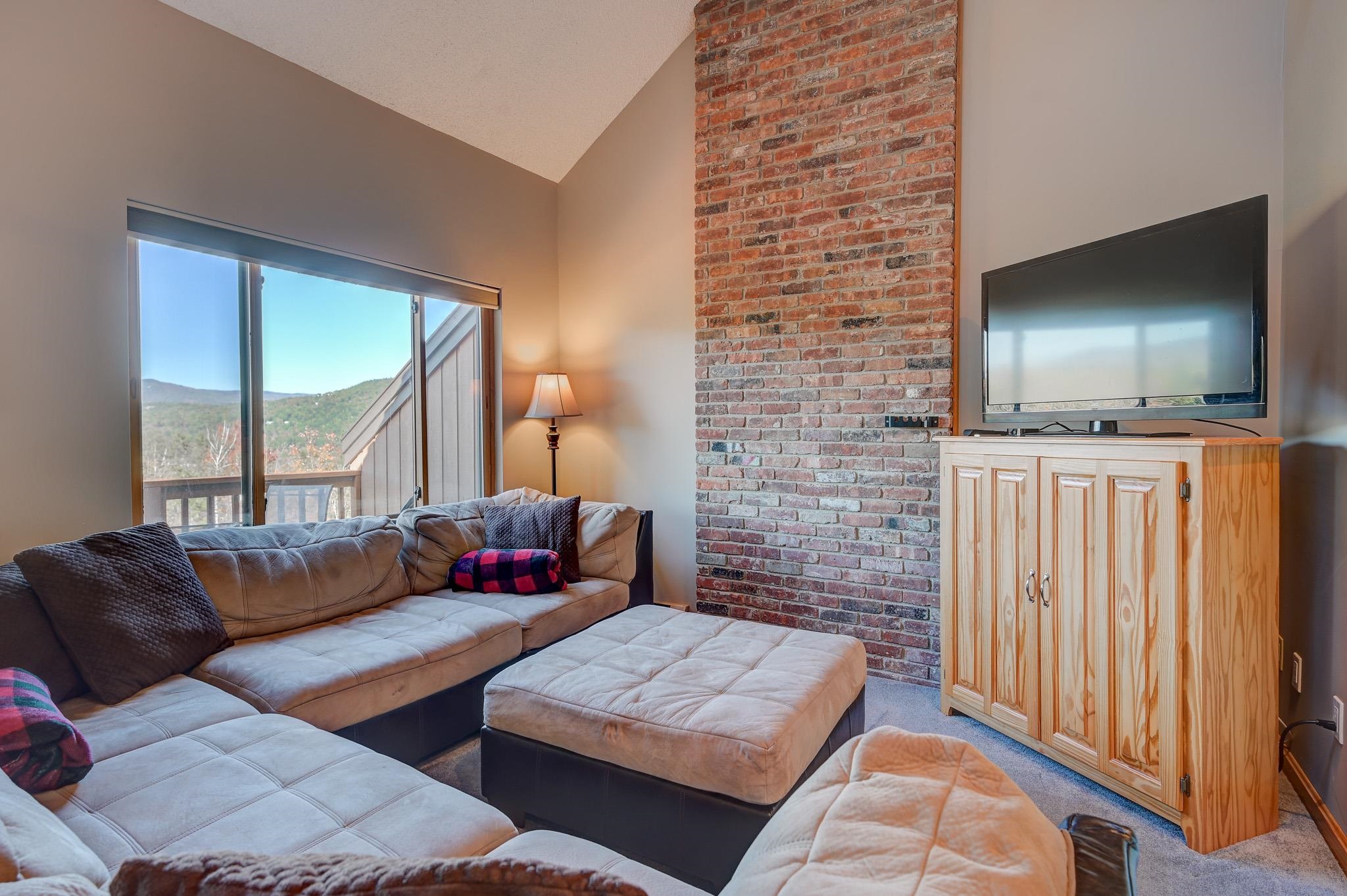 Living room with a sectional, exposed brick wall, and sliding doors overlooking wooded mountains.