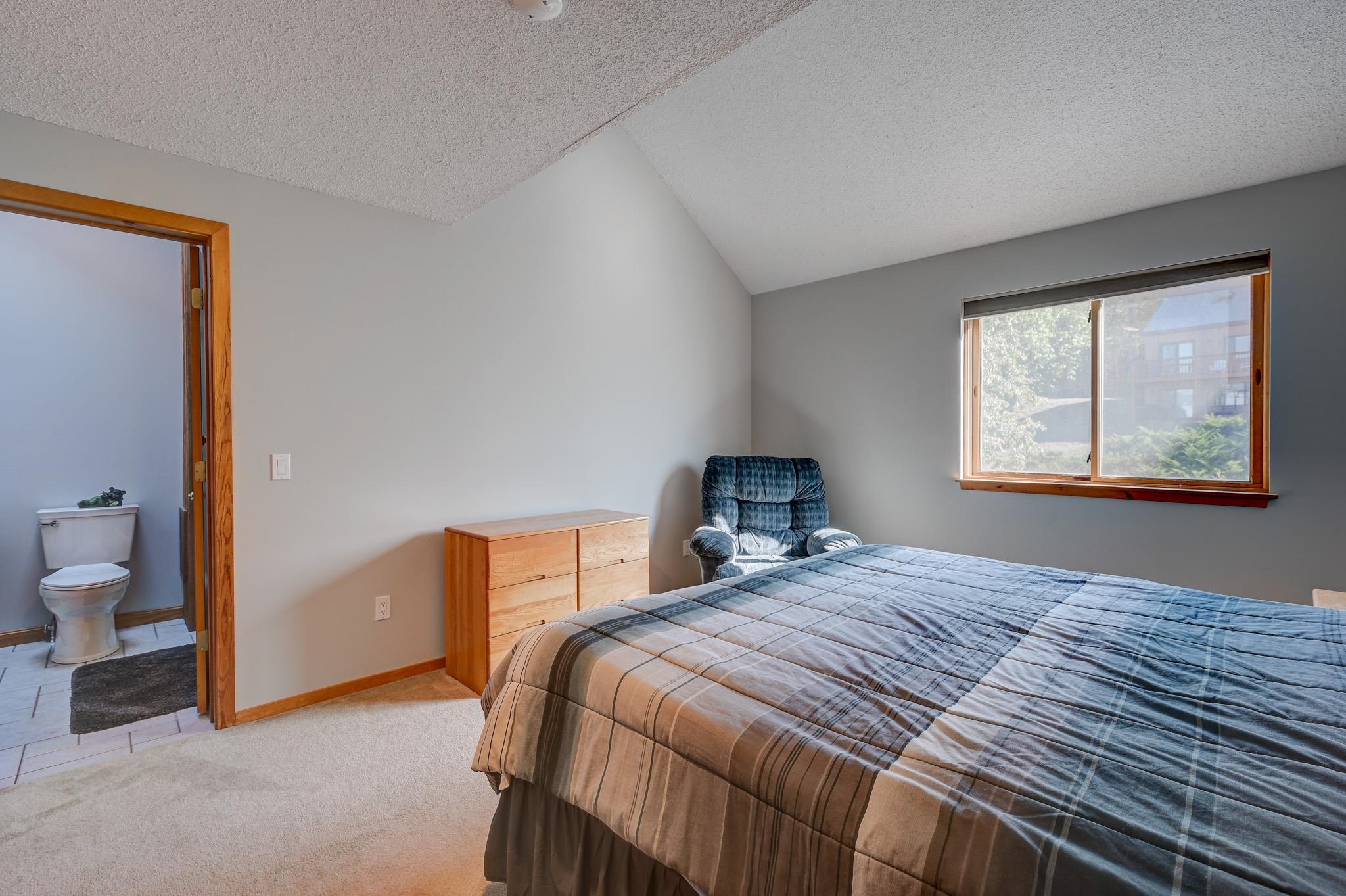 Bedroom with a plaid-covered bed, blue recliner, wooden dresser, and adjoining bathroom.