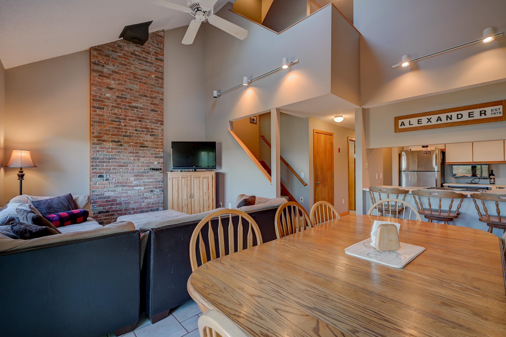 Open-plan living and dining room with a vaulted ceiling, exposed brick wall, and kitchen breakfast bar.