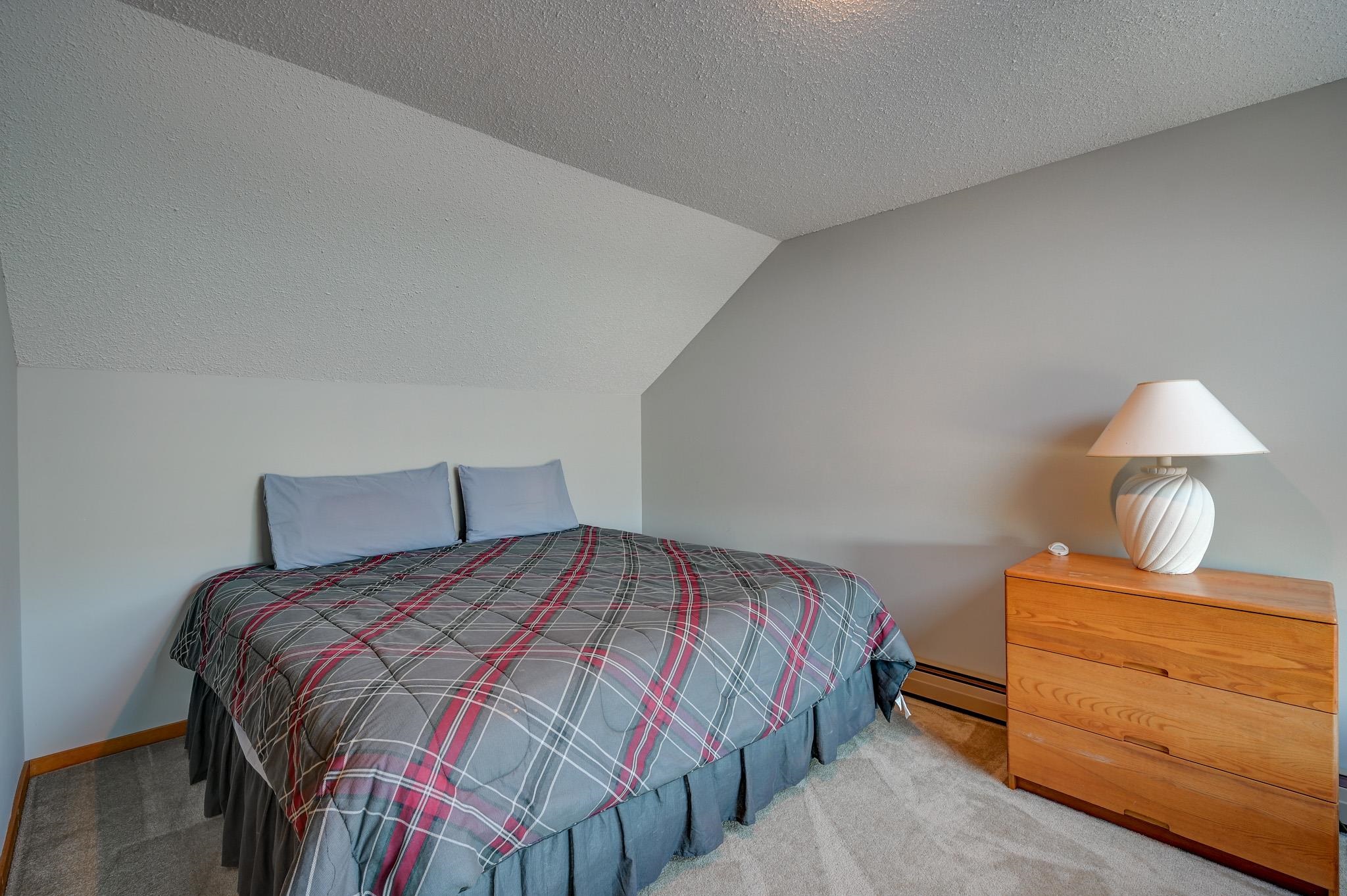 Bedroom with a plaid-covered bed beneath a sloped ceiling and a wooden dresser with a white lamp.