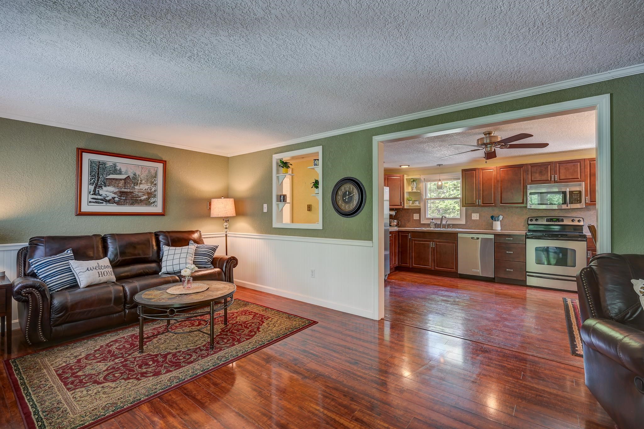 Living room with leather sofas and patterned rug, opening into a kitchen with wood cabinets.