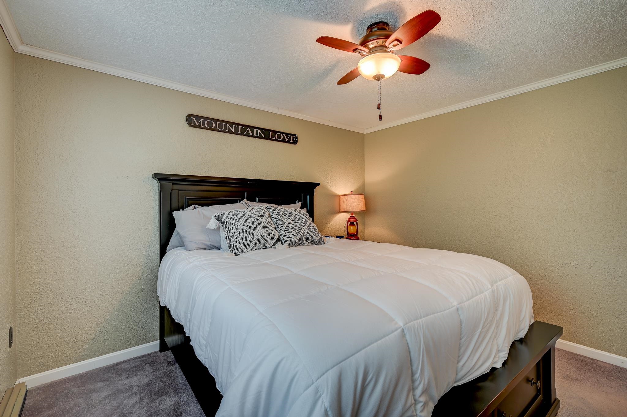 Furnished bedroom with a white bed, dark wood headboard, ceiling fan, and “Mountain Love” sign.