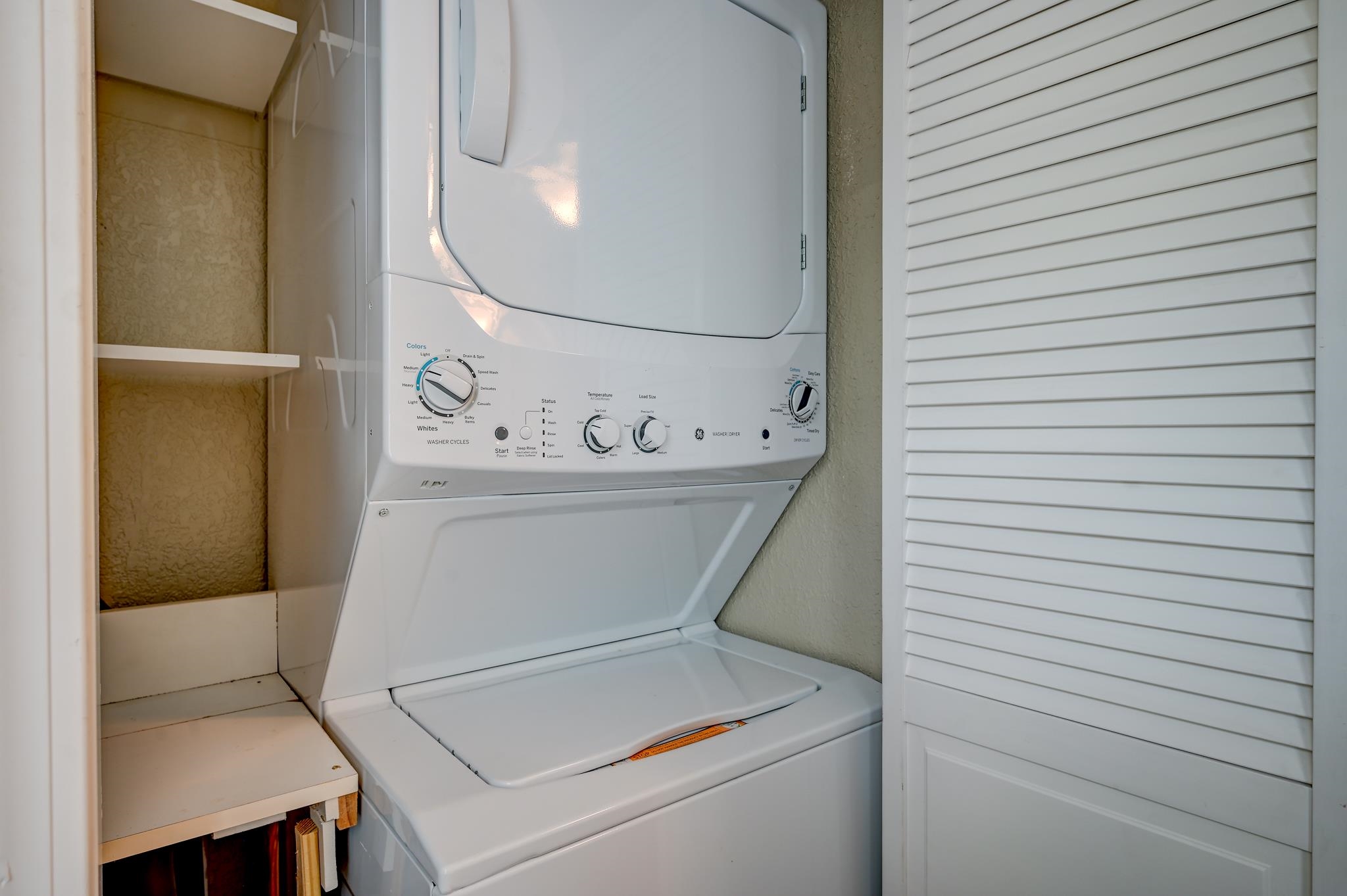 Laundry closet with a stacked white washer and dryer, built-in shelves, and a louvered door.