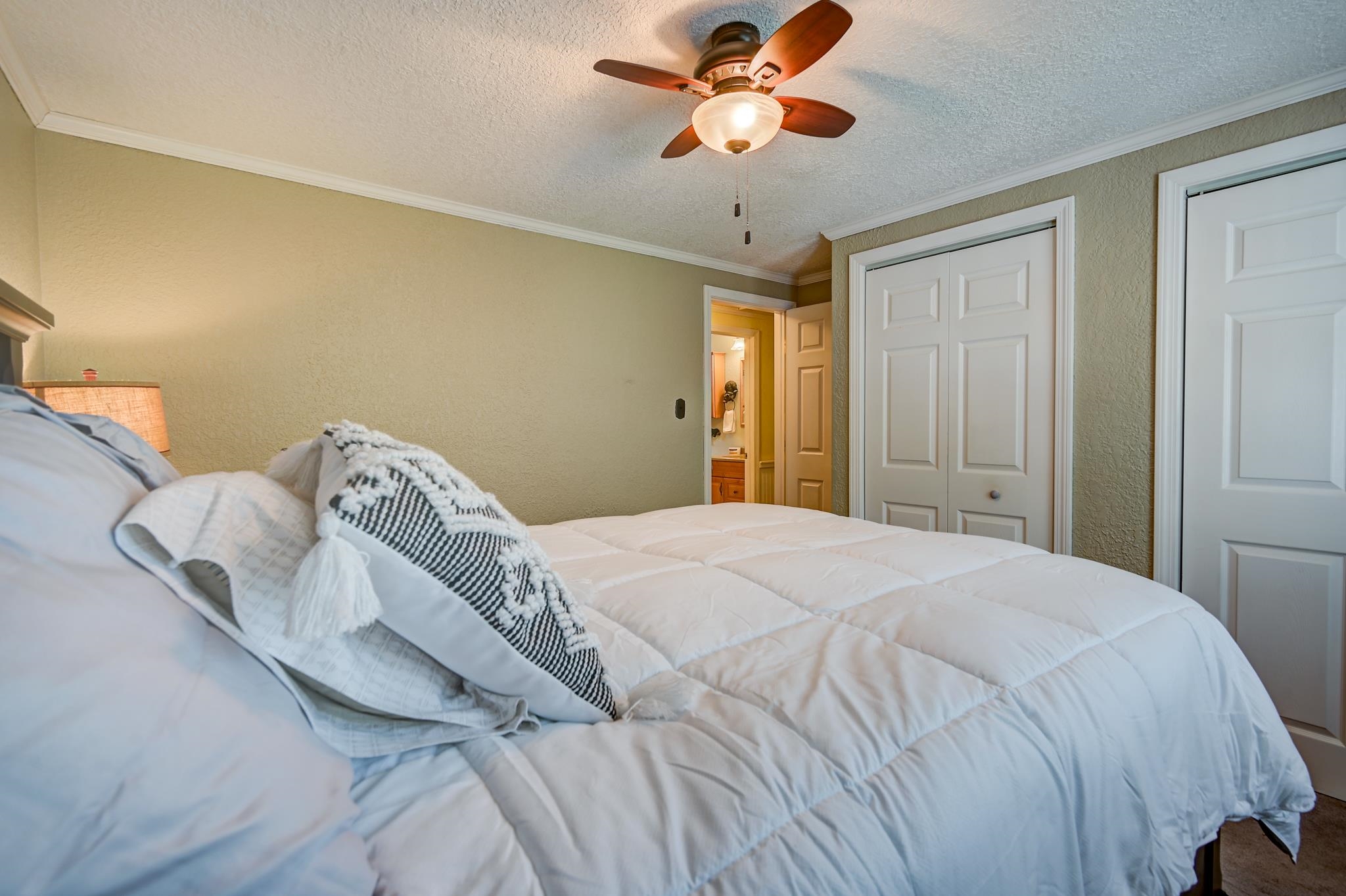 Bedroom with a white quilted bed, ceiling fan, and open doorway to a bathroom.