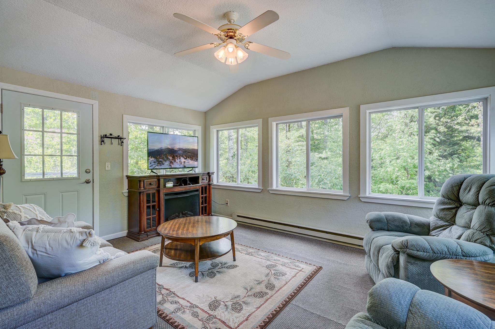 Furnished living room with vaulted ceiling, gray seating, and large windows overlooking leafy trees.