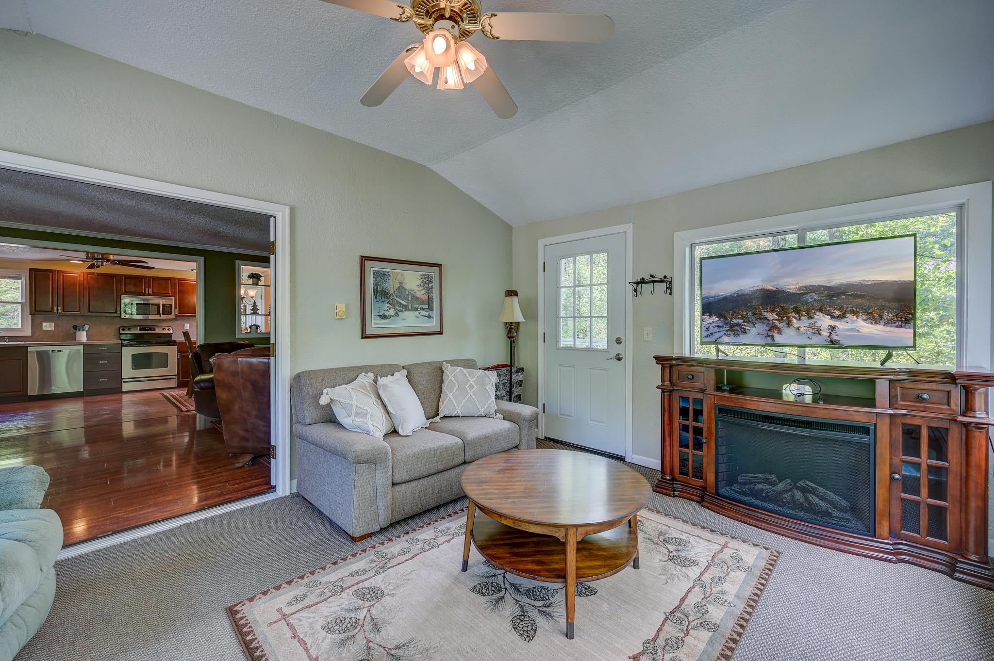 Living room with vaulted ceiling, gray sofa, wooden fireplace console, and an adjoining kitchen.