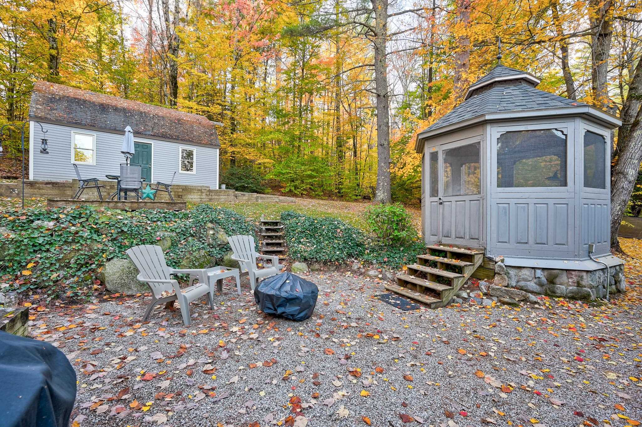 Gravel patio with a screened gazebo, outdoor chairs, and a small house among autumn trees.