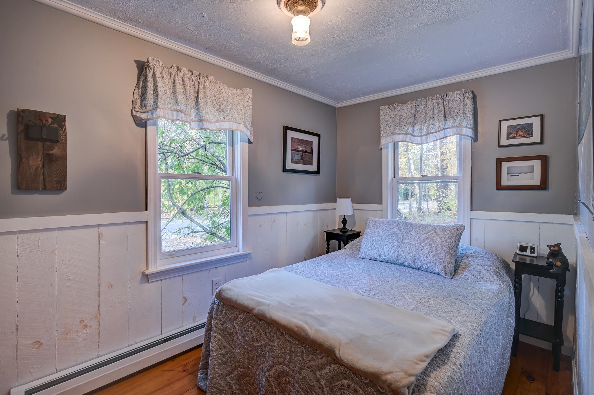 Furnished bedroom with a patterned bed, two curtained windows, and white wainscoting.