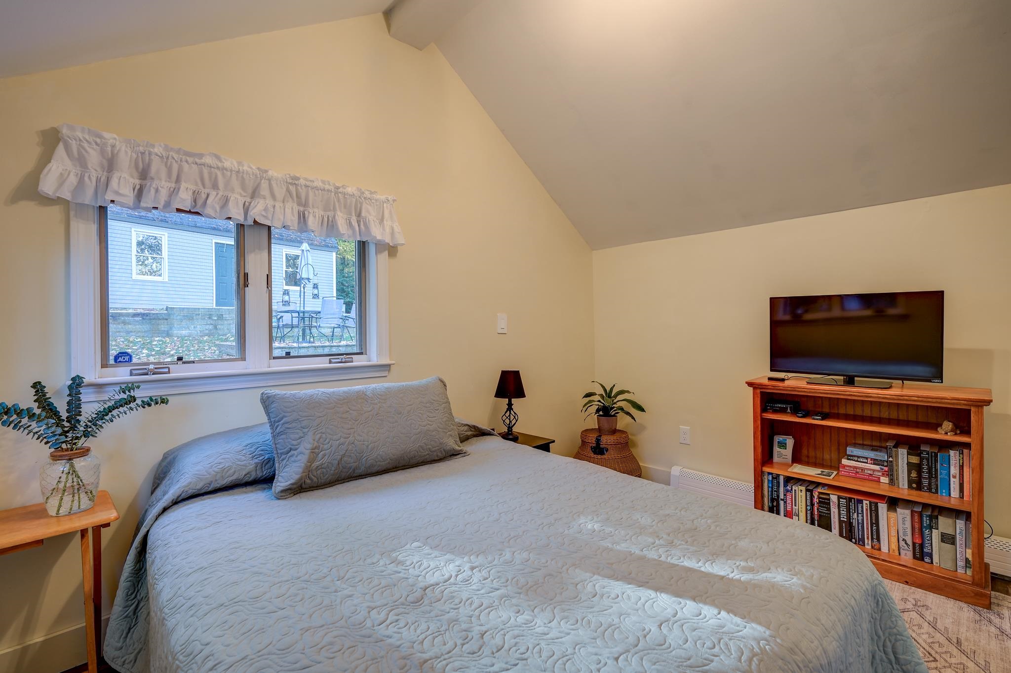 Furnished bedroom with a large bed, sloped ceiling, wide window, and television on a wooden bookcase.