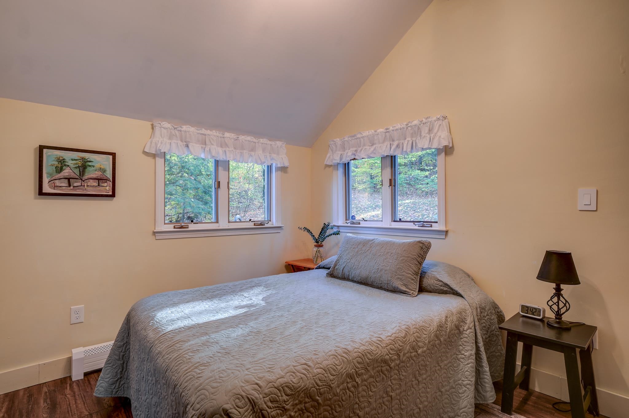 Bedroom with a gray quilted bed beneath a sloped ceiling and windows overlooking leafy trees.