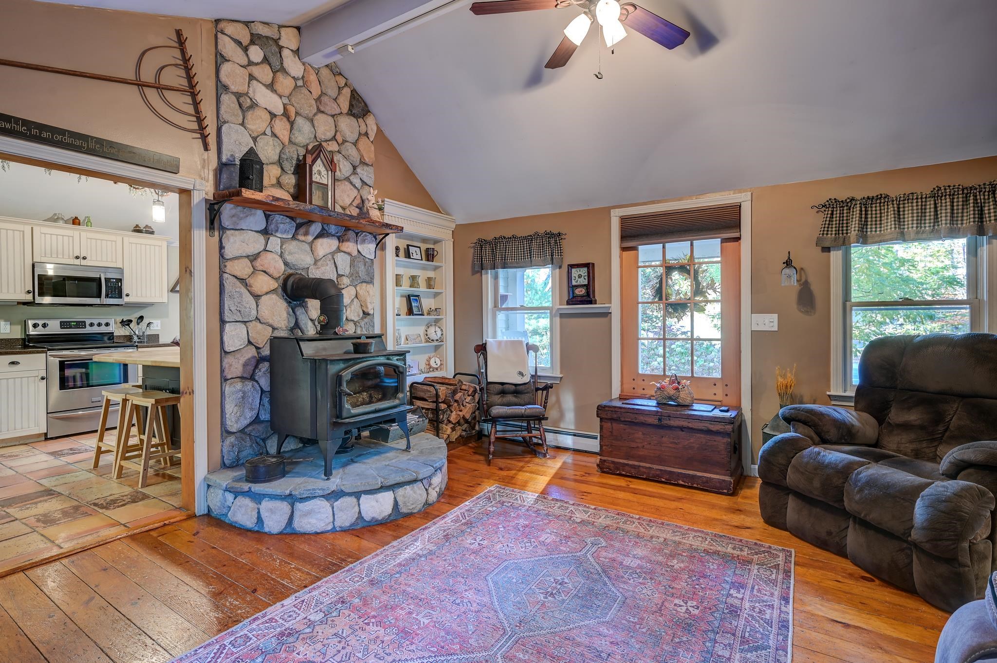 Living room with a stone fireplace and wood-burning stove, hardwood floors, and a view into the kitchen.