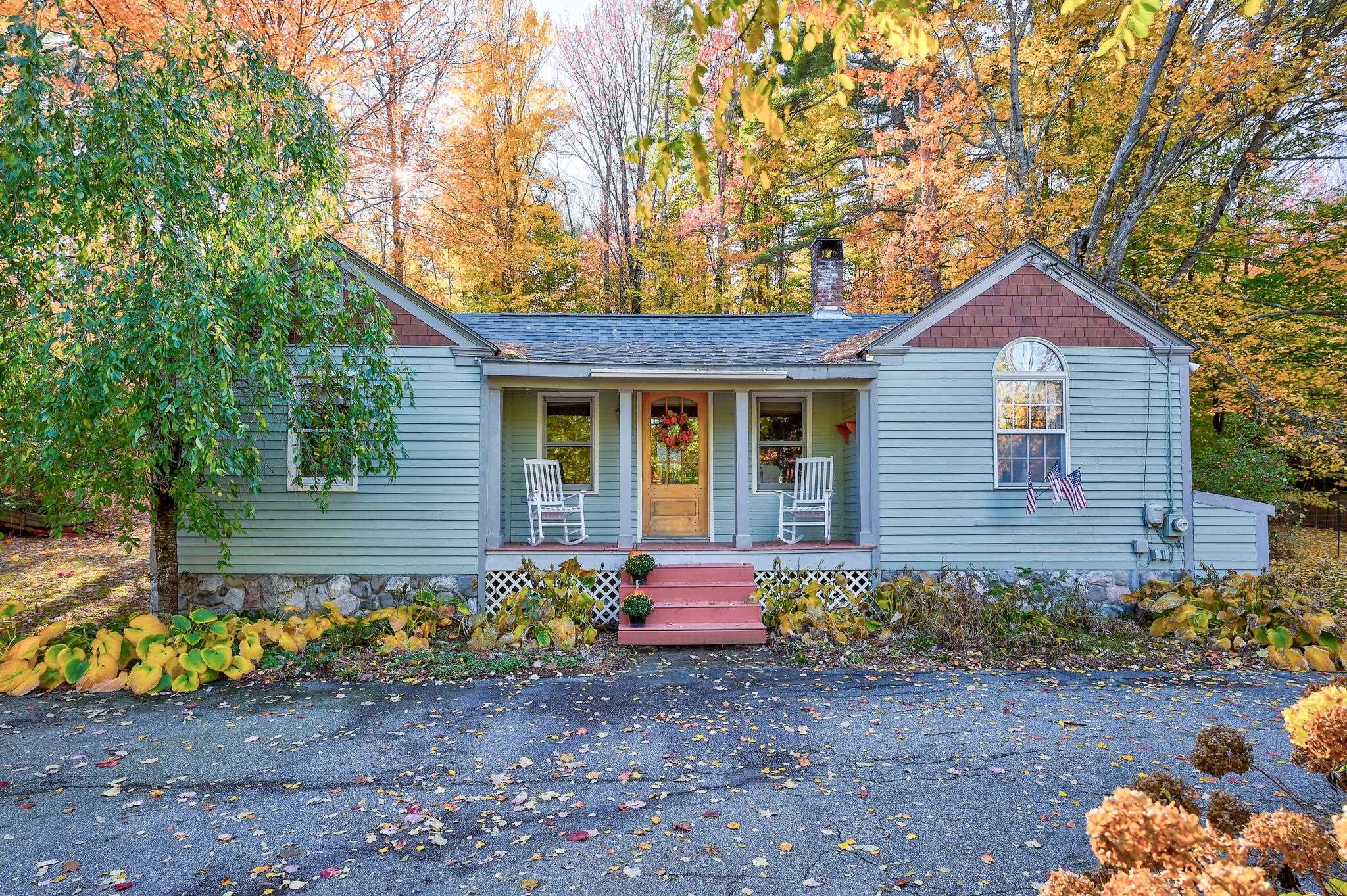 Pale blue house with red front steps and white rocking chairs, framed by autumn foliage.