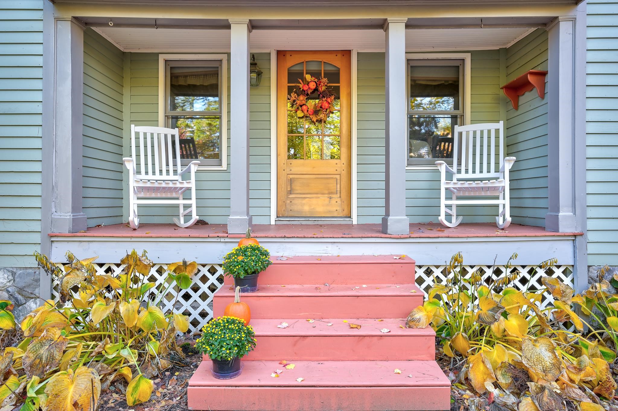 Covered front porch with white rocking chairs, autumn wreath, and pink steps decorated with pumpkins and flowers.
