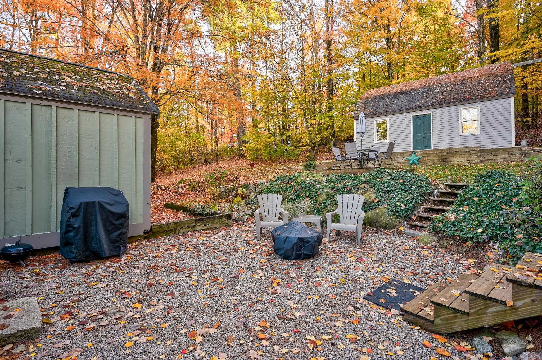 Gravel patio with Adirondack chairs and a covered fire pit beside a small outbuilding in autumn woods