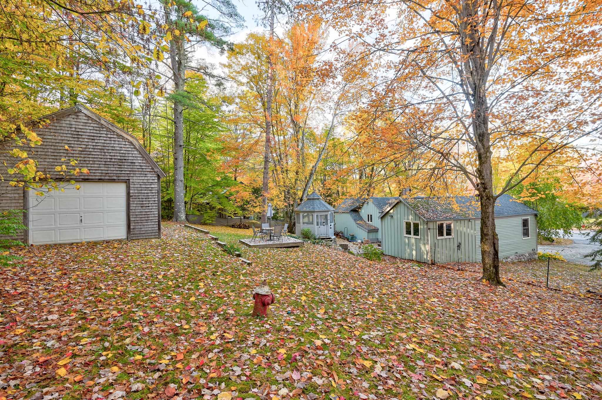 Light-green house and detached garage in a leafy autumn yard, with a gazebo, patio, and water nearby.