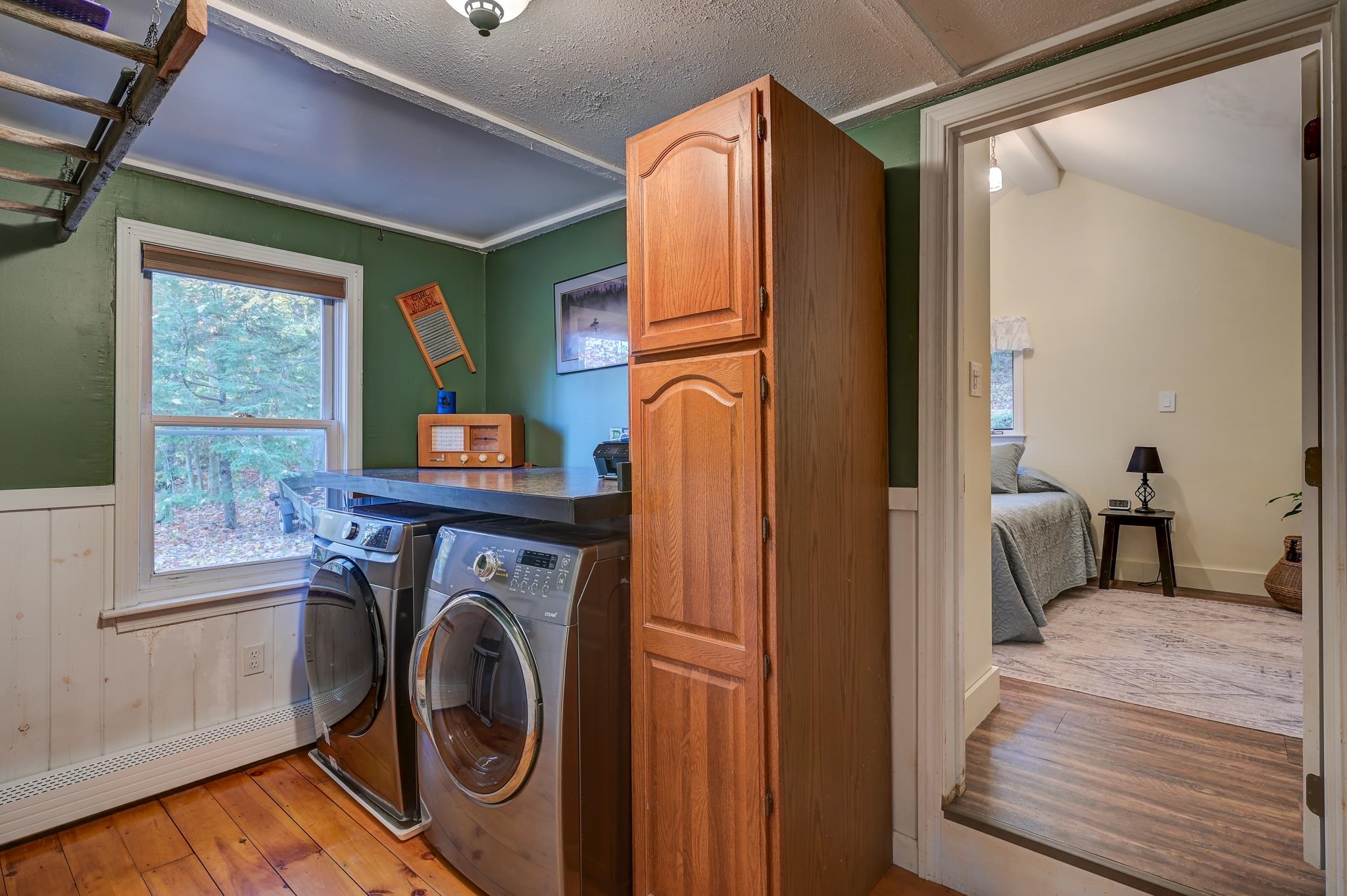 Laundry room with front-loading washer and dryer, wooden storage cabinet, and doorway to a bedroom.
