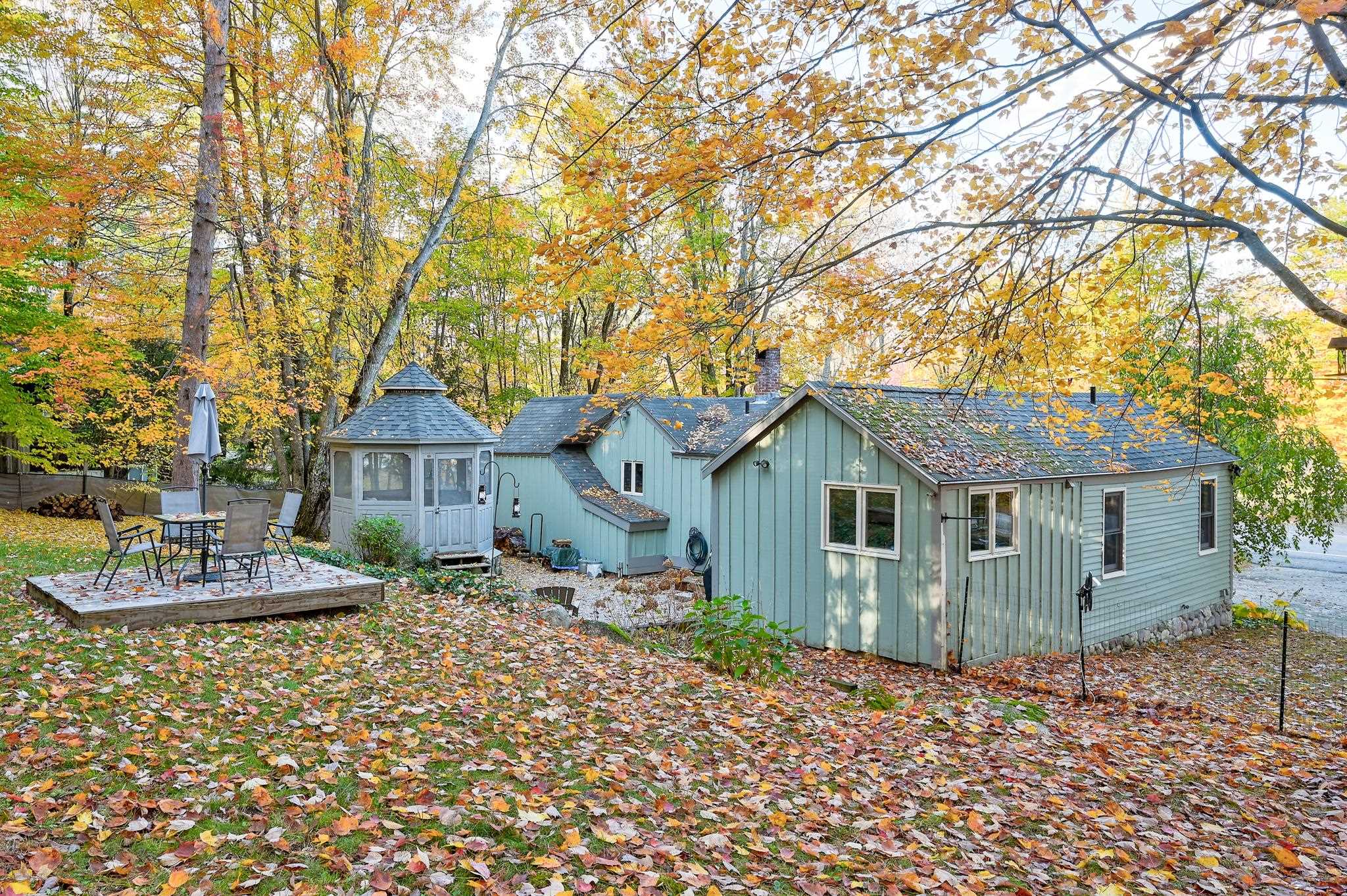 Light blue-gray house with an enclosed gazebo and outdoor seating deck beneath autumn trees.