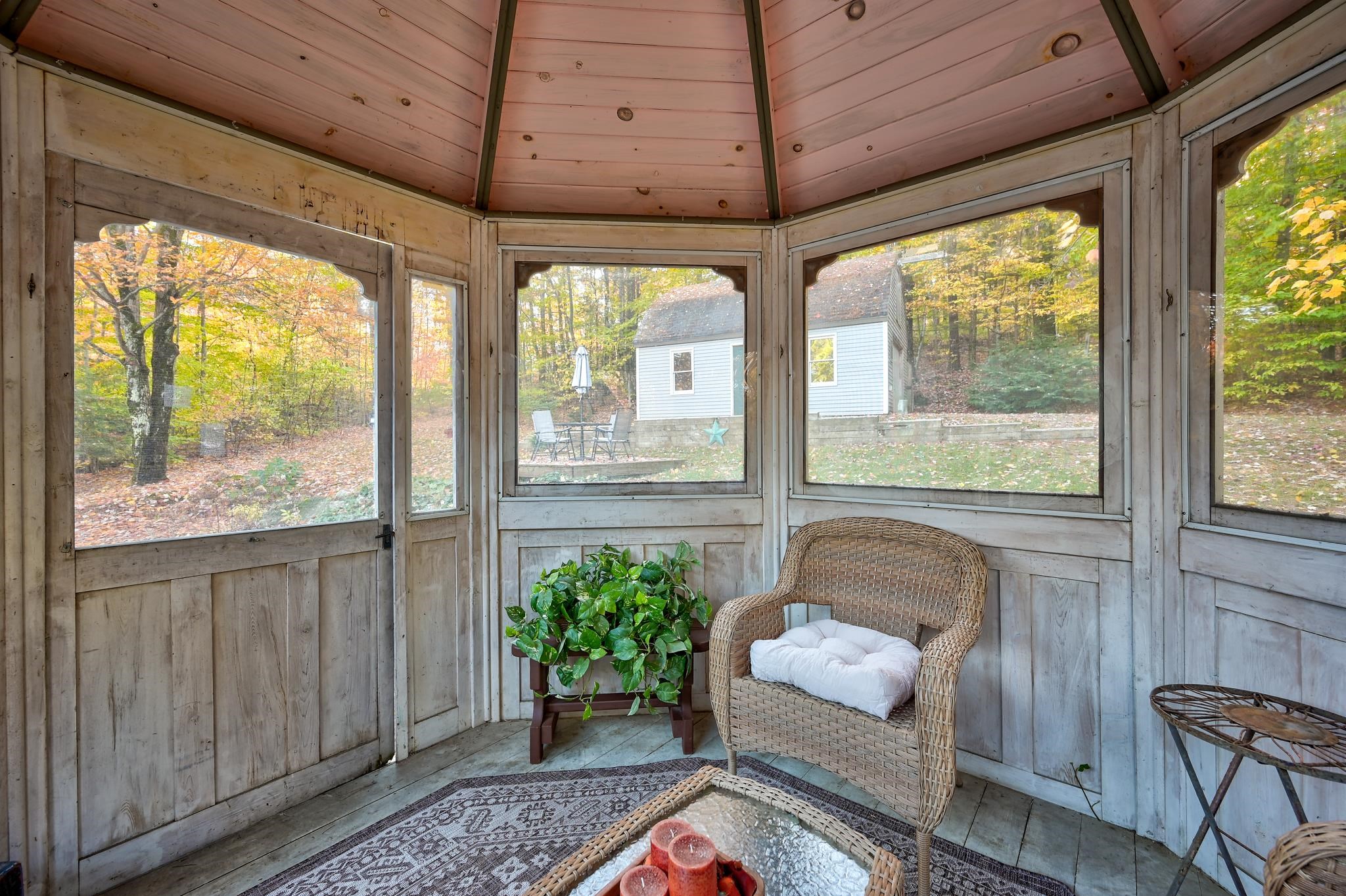 Enclosed sunroom with a wicker chair and plant, overlooking autumn trees and a small outbuilding.