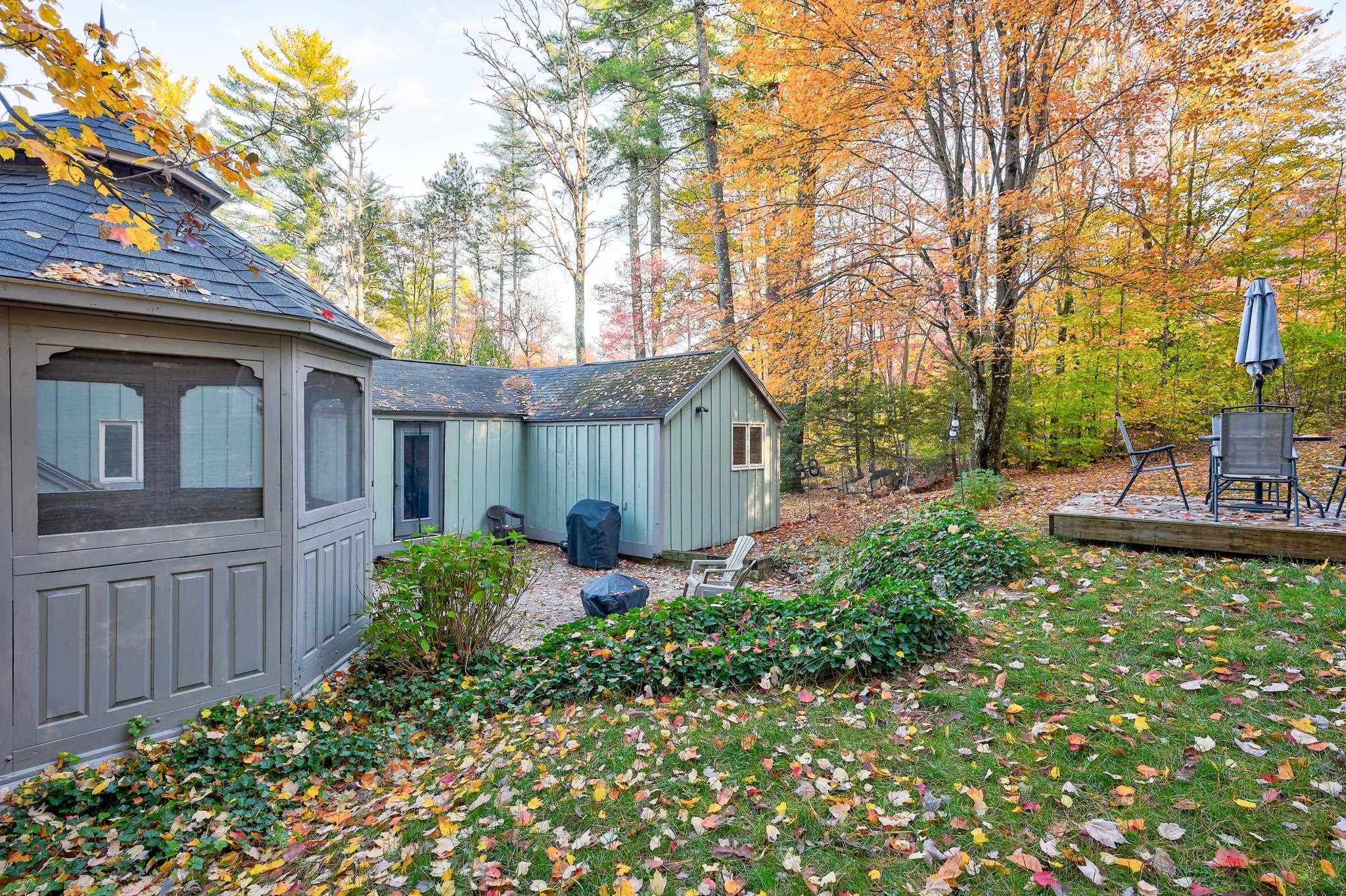 Wooded backyard with a screened gazebo, pale green outbuilding, and raised deck amid autumn foliage.