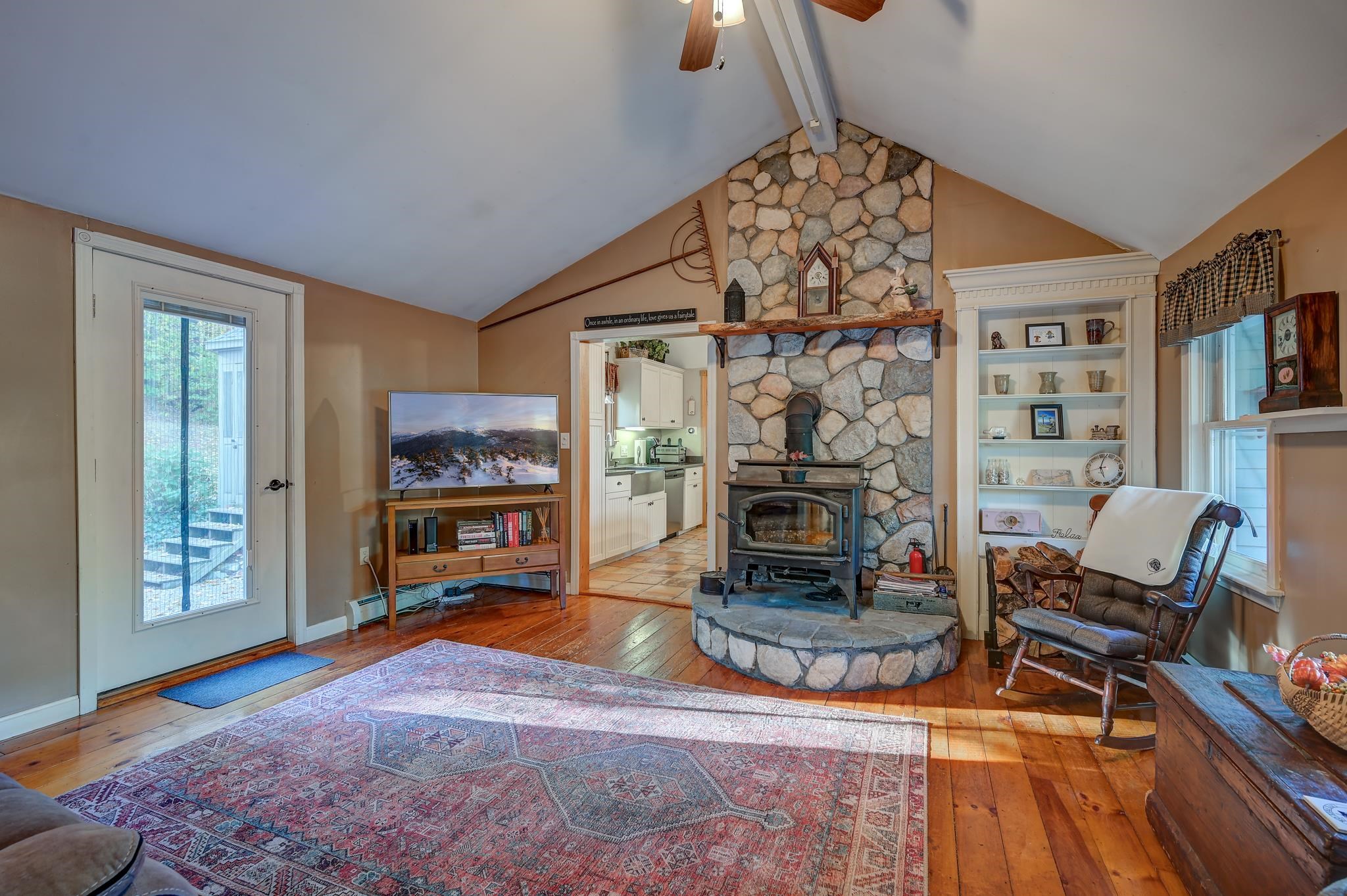 Furnished living room with a vaulted ceiling, stone fireplace, wood stove, and hardwood floors.